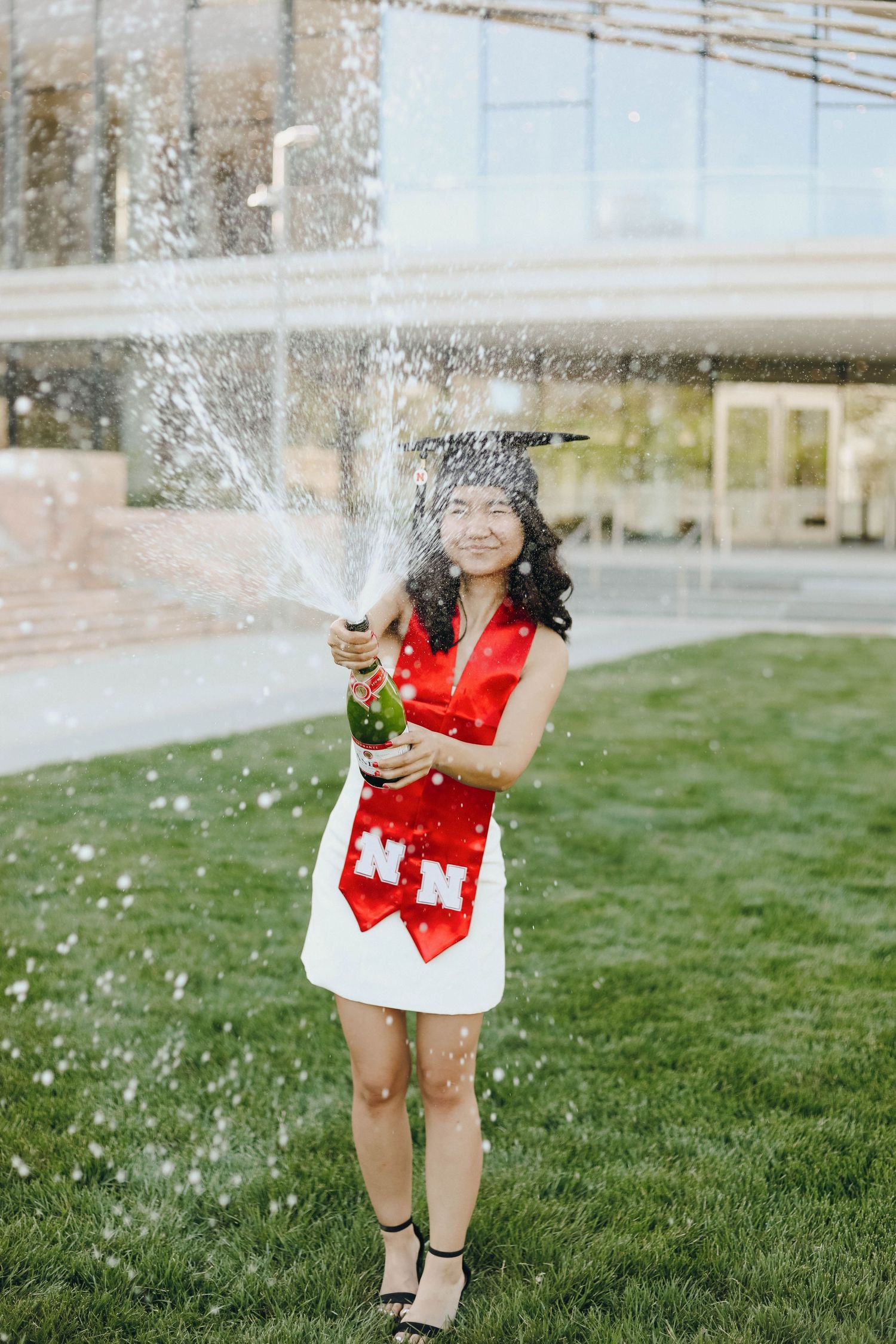 student celebrating graduation with a cap and gown spraying a bottle of finger lakes sparkling wine