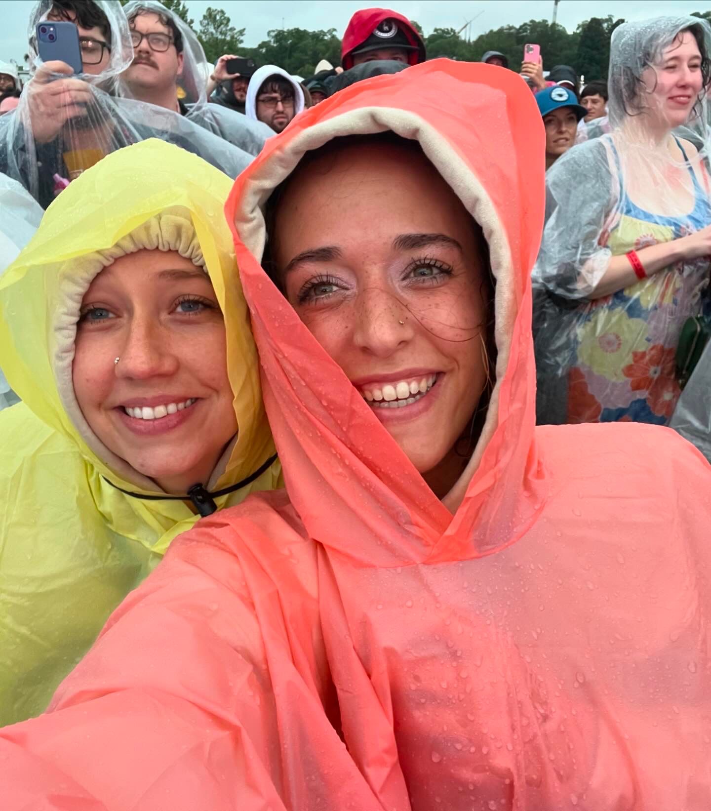 Two smiling women wearing colorful rain ponchos at an outdoor festival in the rain, taking a selfie.