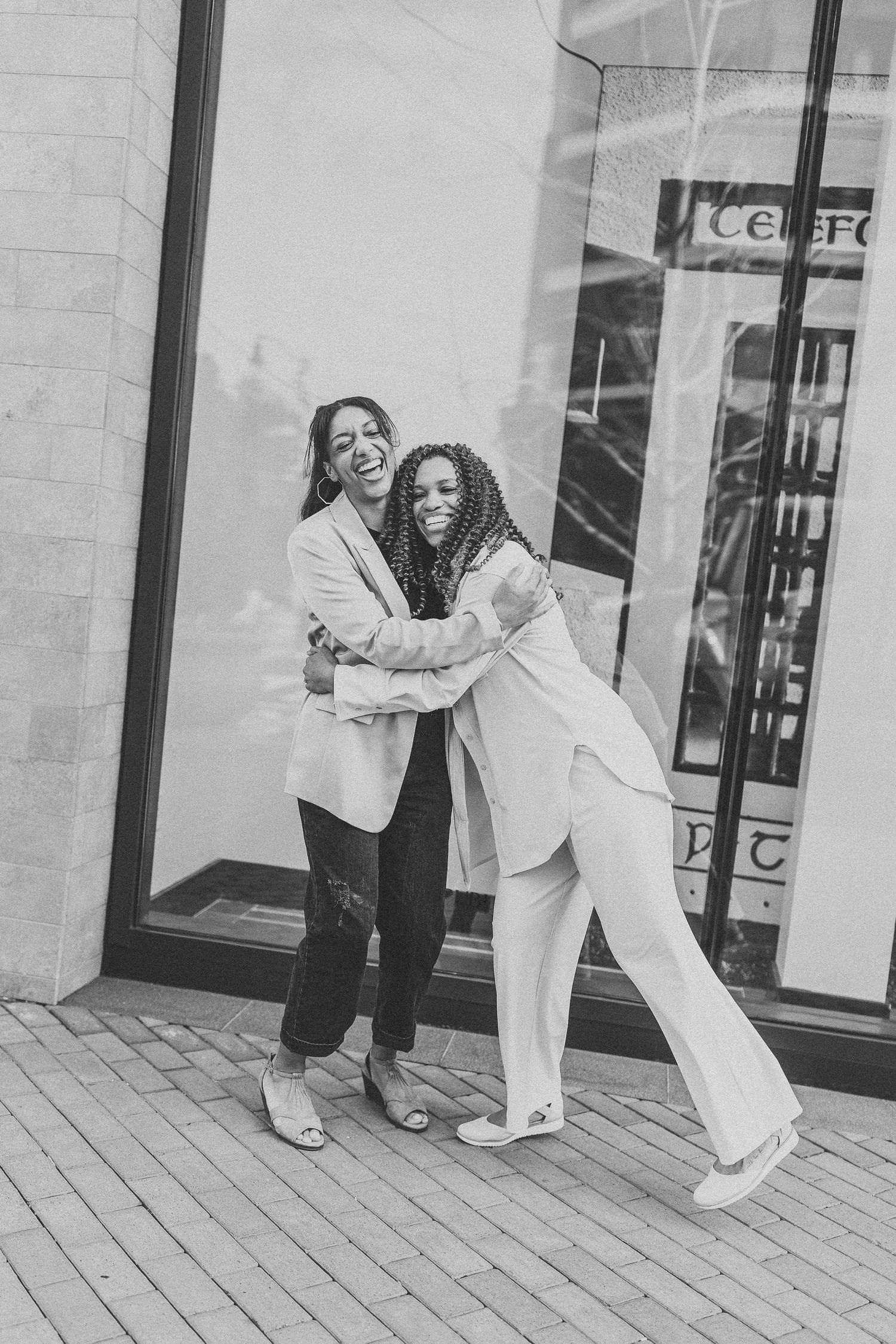 Two women hugging and laughing together outside a building in black and white photo.