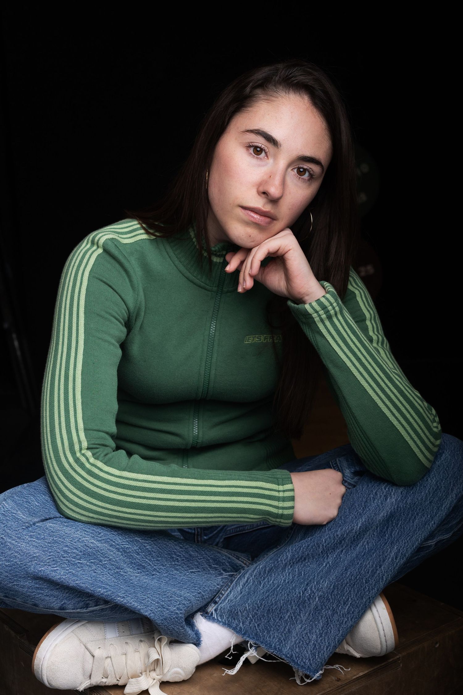 Young brunette woman in green Adidas track jacket and jeans sitting cross-legged on floor.