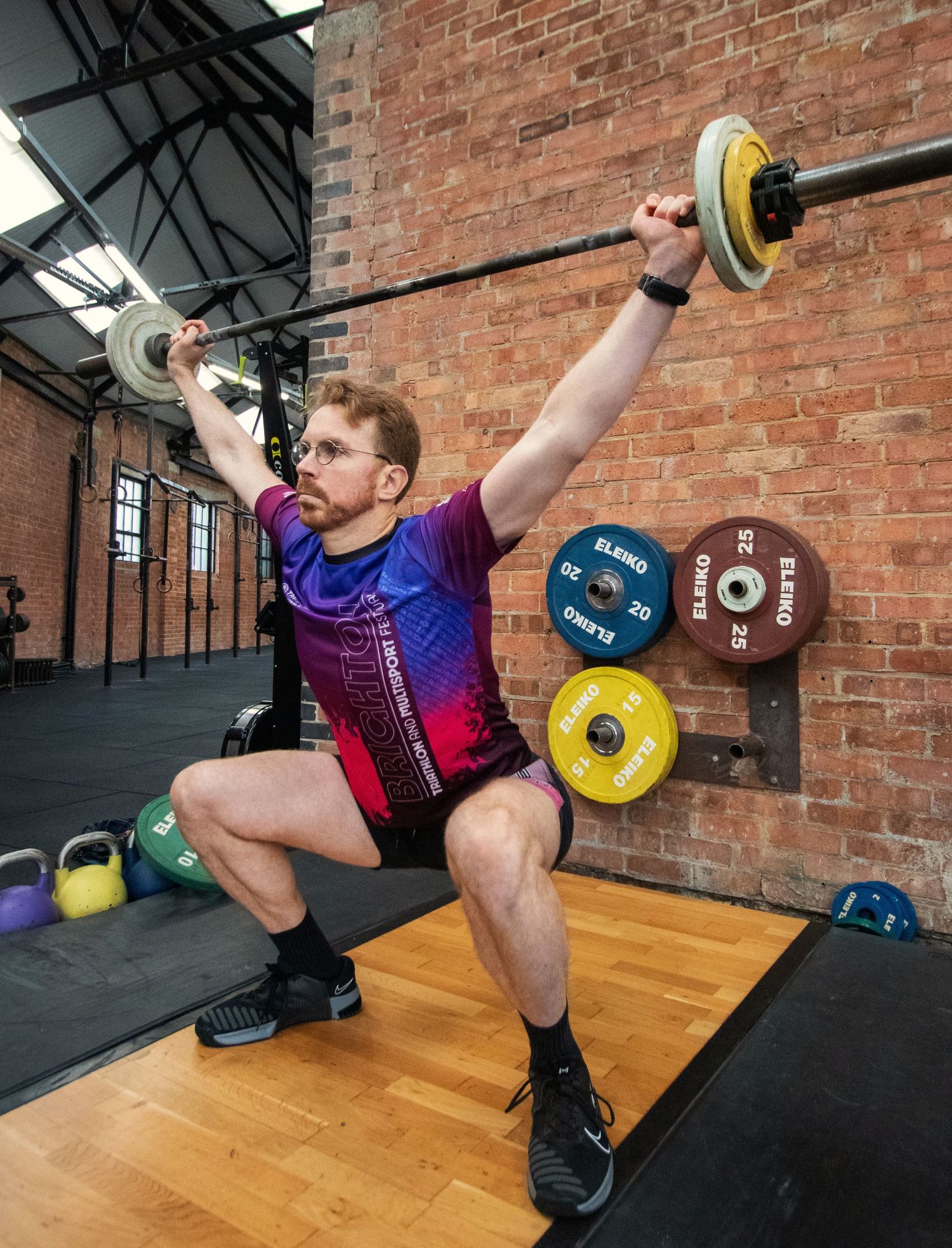 Male athlete performing an overhead squat with barbell in a brick-wall CrossFit gym.
