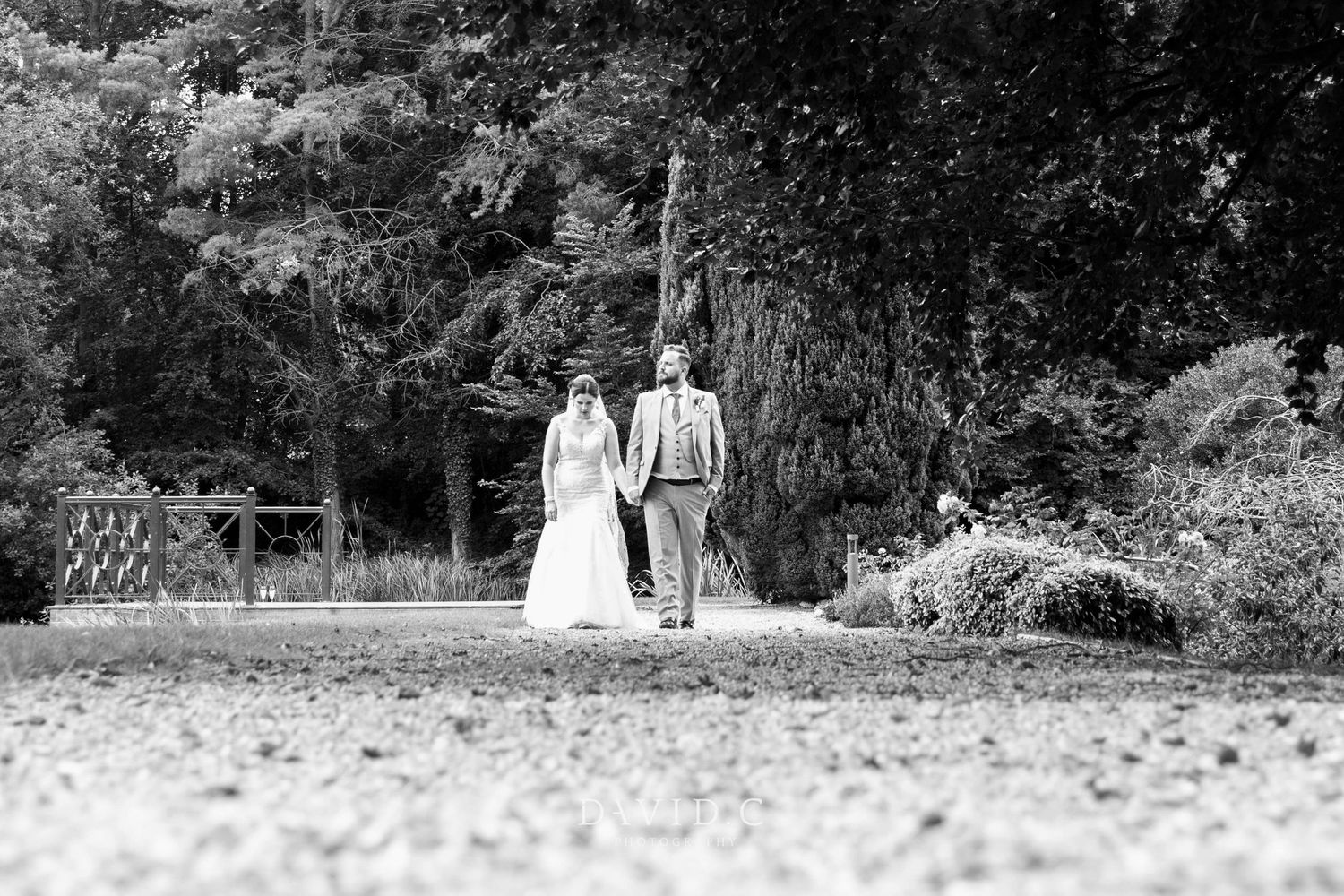 Black and white photo of bride and groom walking hand in hand through lush green park grounds.