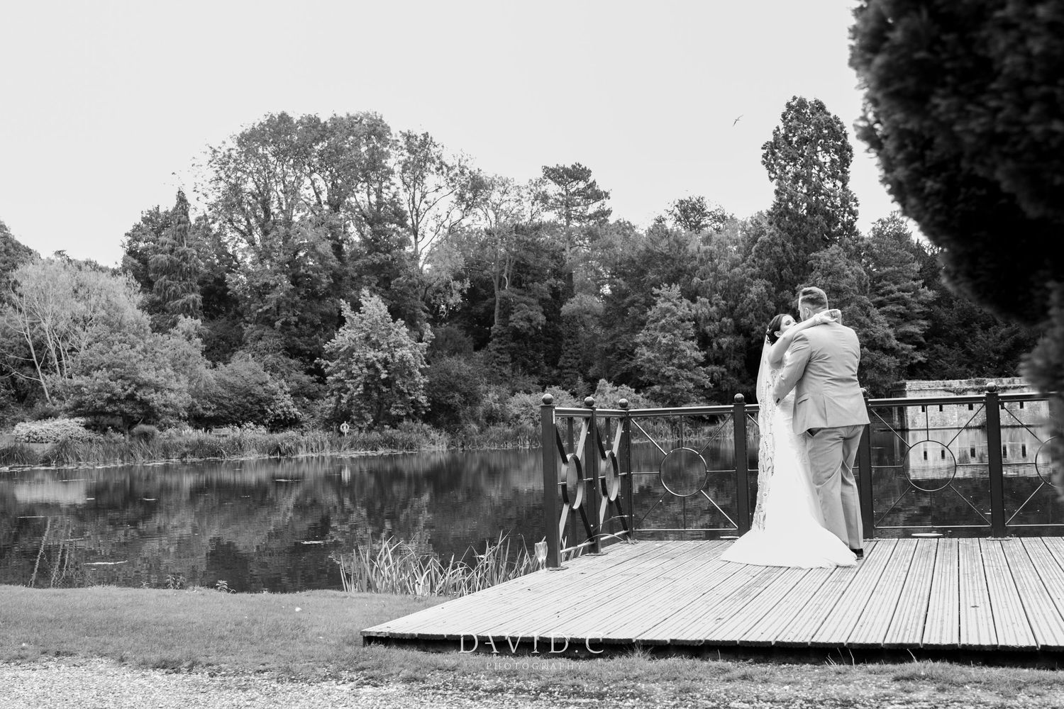Wedding Photography - Black and white photo of bride and groom embracing on wooden dock by tranquil lake surrounded by trees.