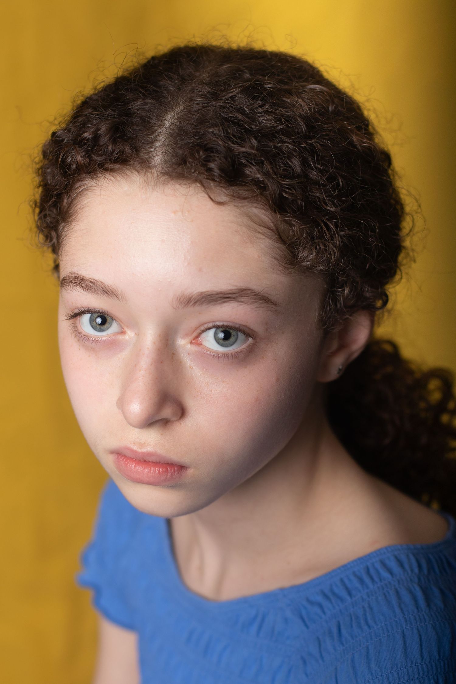 Young girl with dark curly hair and blue eyes against yellow background wearing blue top.