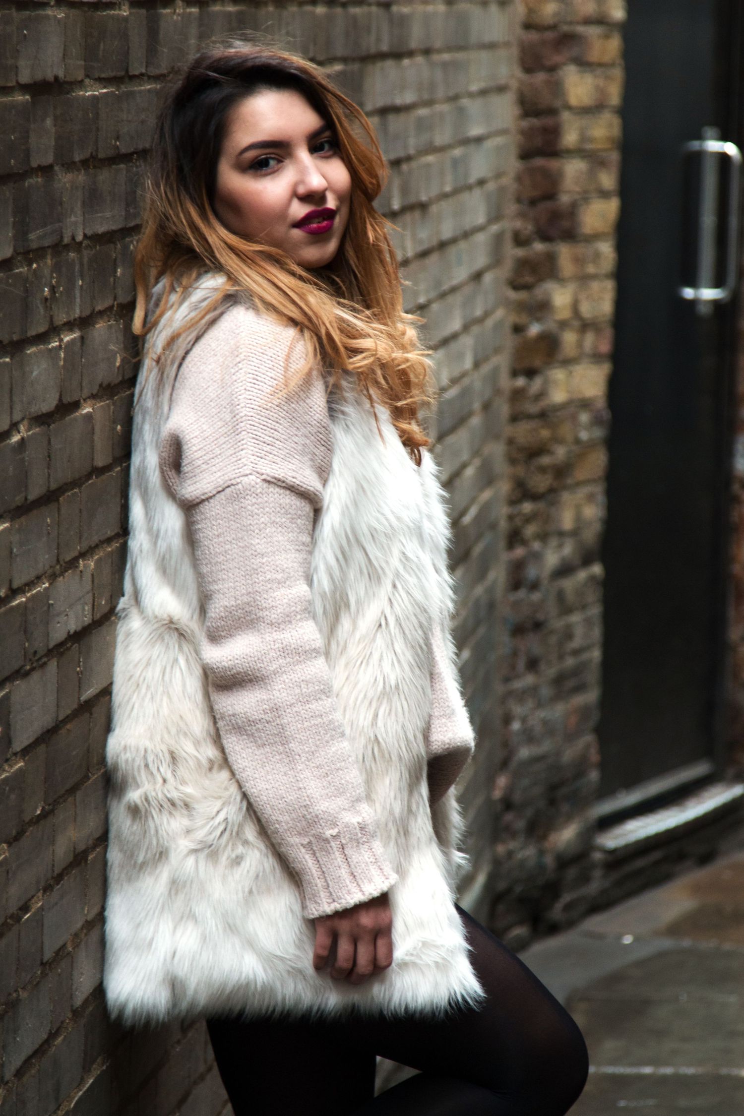 Young woman in white faux fur vest and pink knit sweater posing against brick wall outdoors.