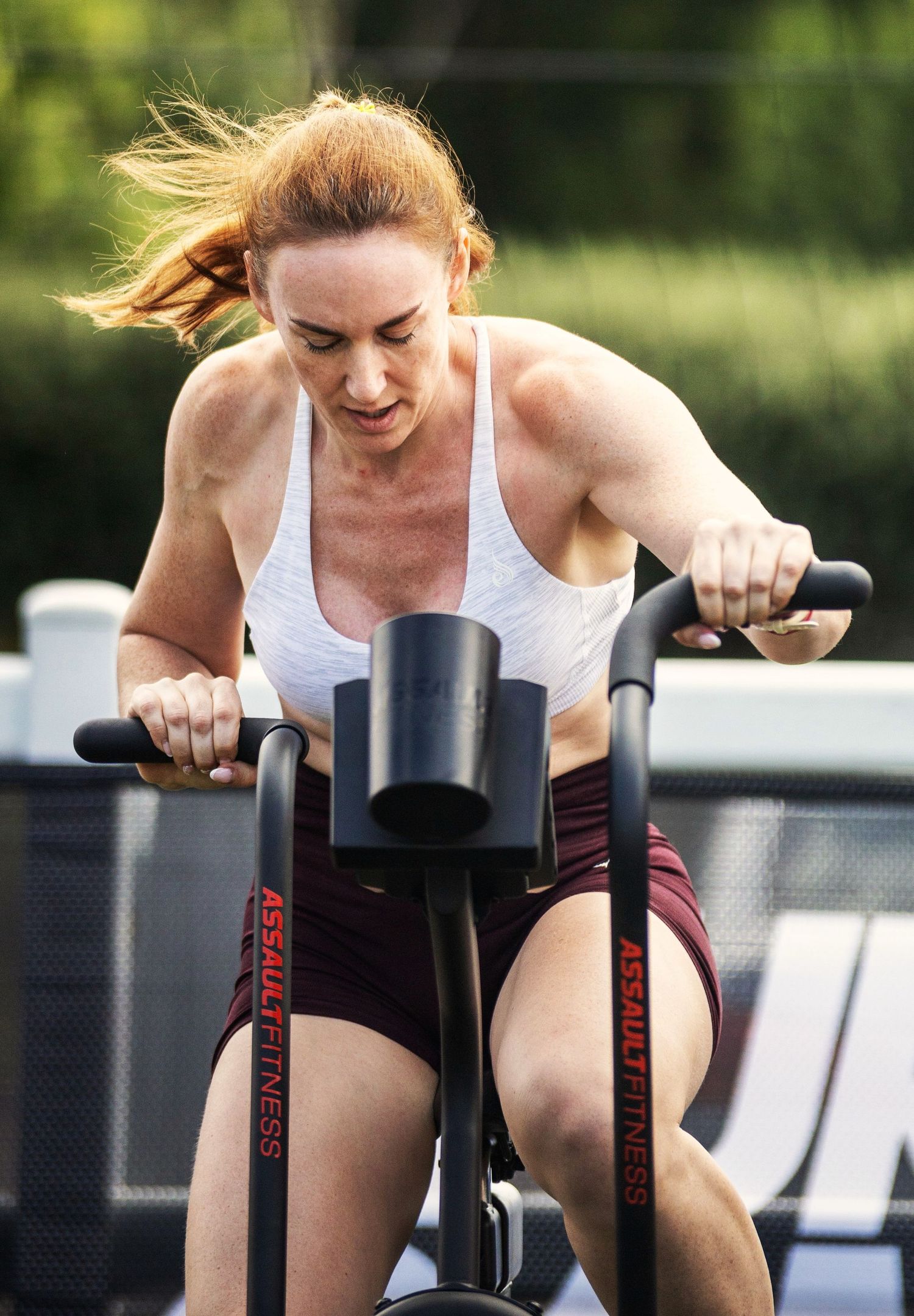Woman with red hair intensely cycling on an Assault Fitness bike outdoors.