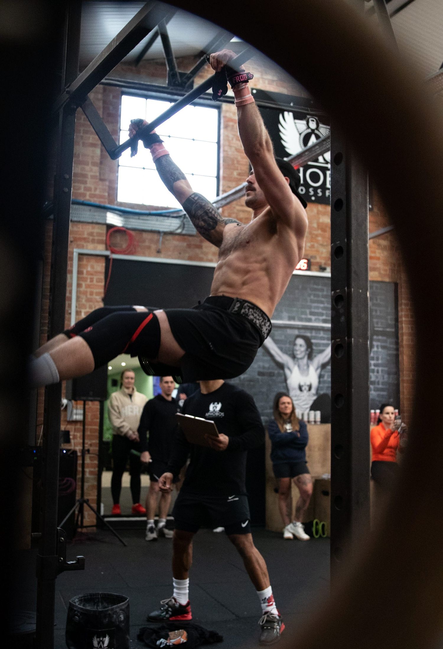 Shirtless tattooed male athlete performing bar muscle-ups in a CrossFit gym.