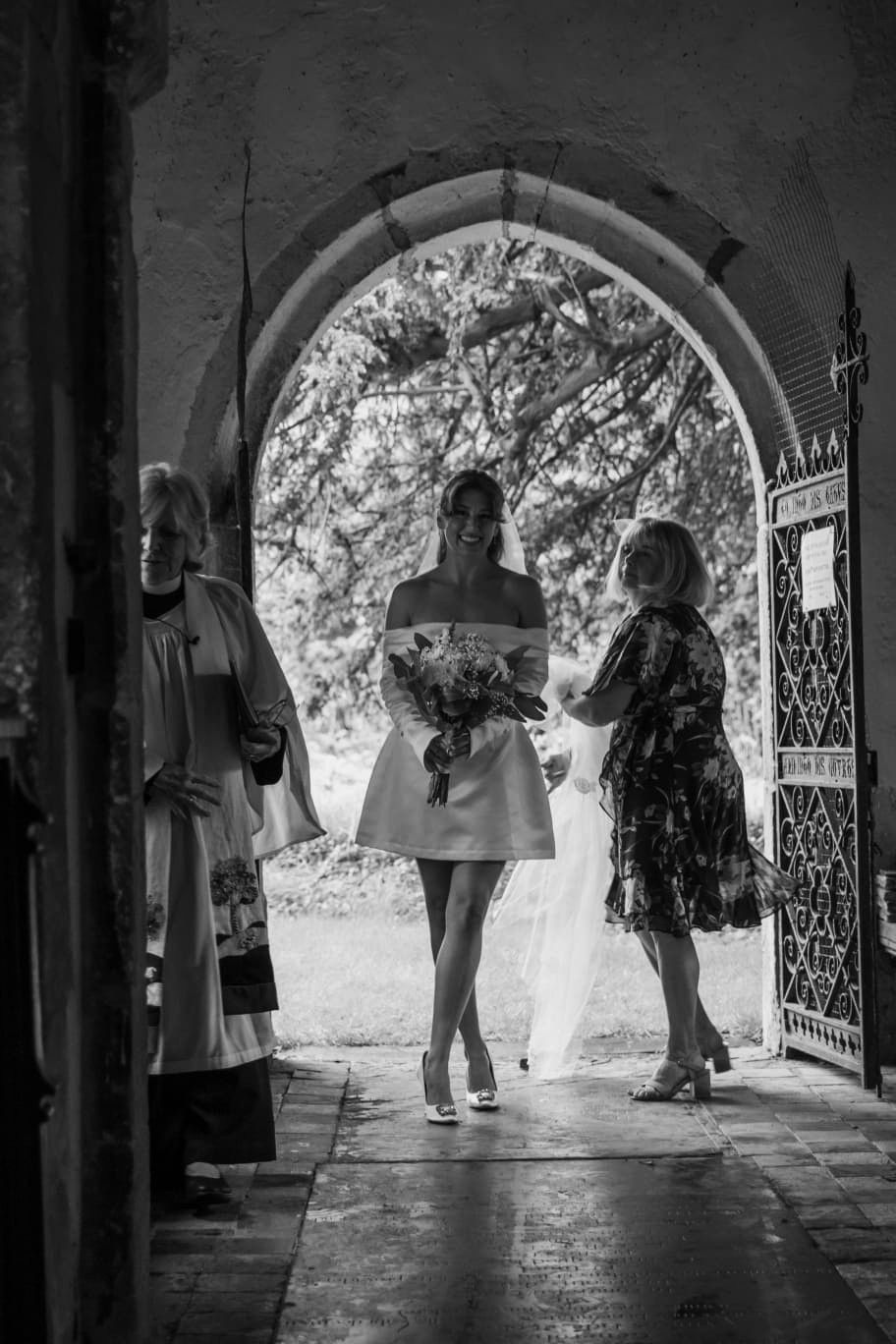 Black and white photo of a bride in short dress holding bouquet at church entrance with officiant and guest.