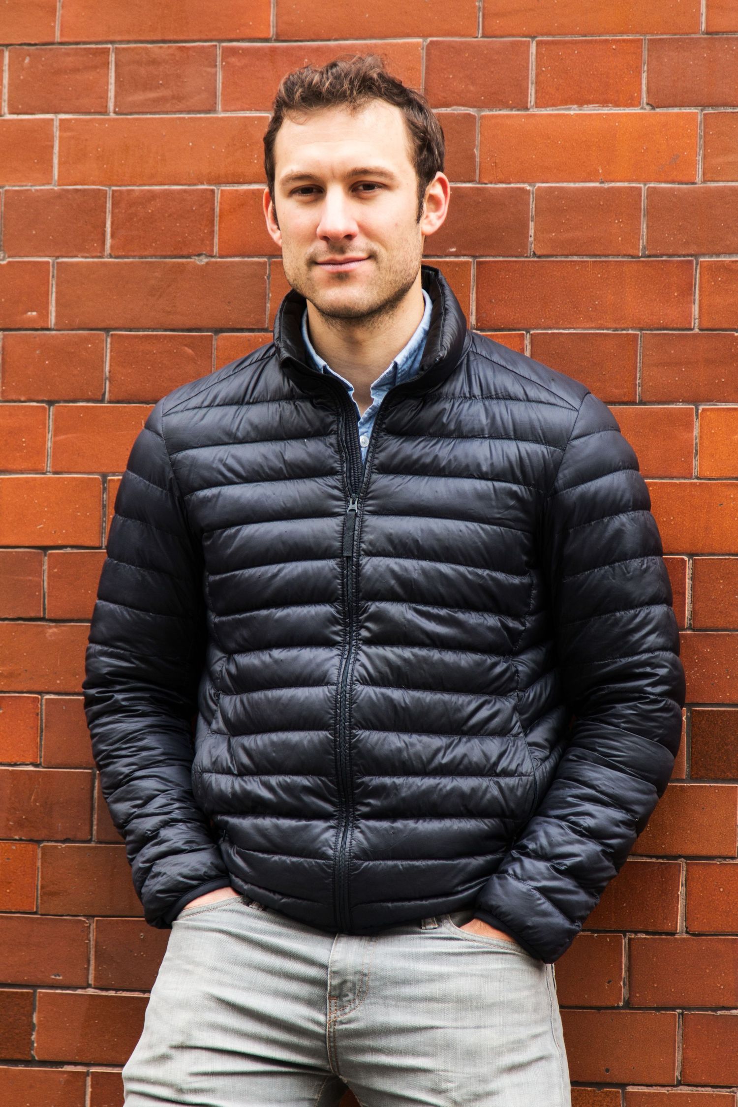 Young man in black puffer jacket standing against red brick wall smiling.