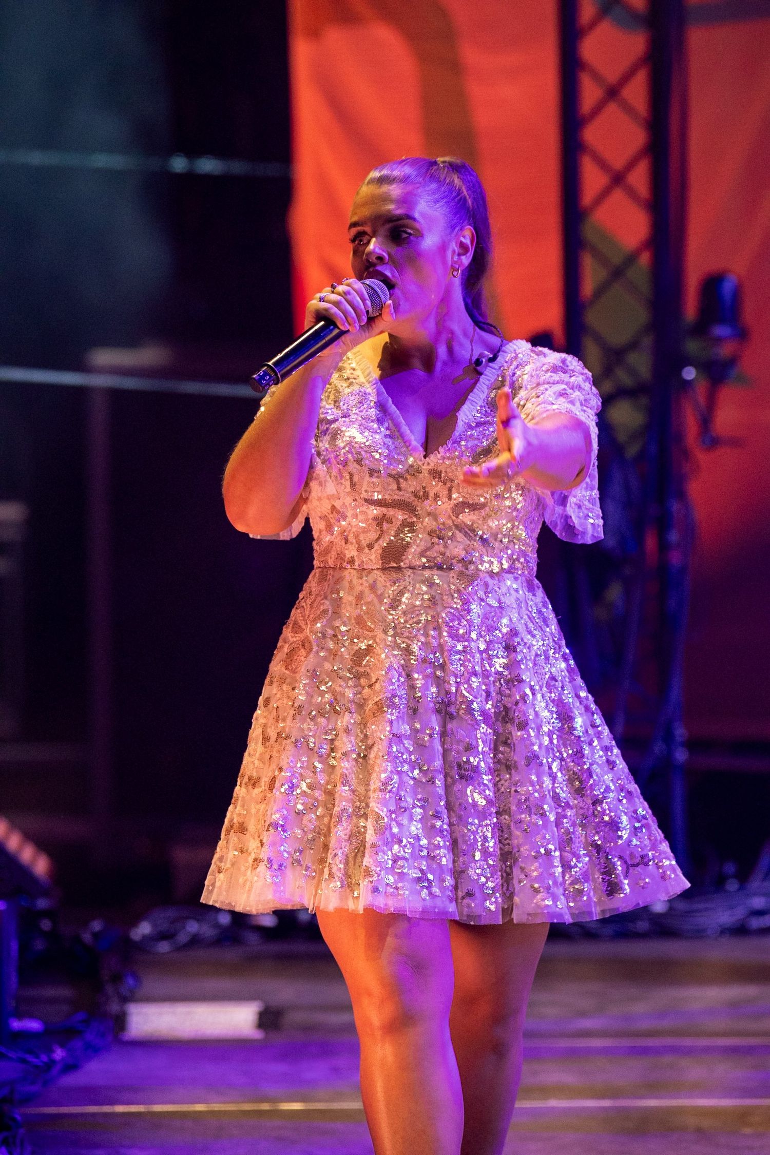 Female singer in sparkly silver dress performing with microphone on a purple-lit stage.