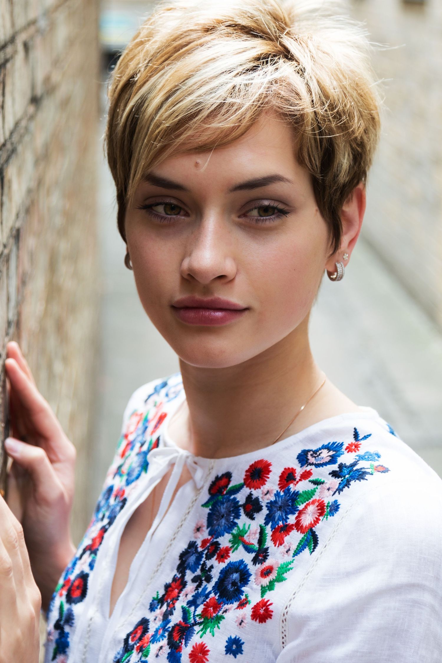 Portrait photography - Young woman with short blonde pixie cut in floral embroidered white top near stone wall.