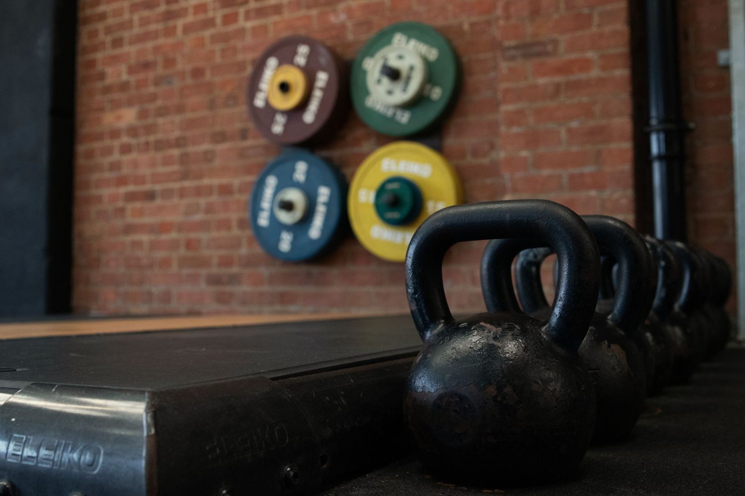 Fitness Photography - Black cast iron kettlebells lined up on a rack with colorful weight plates on brick wall.