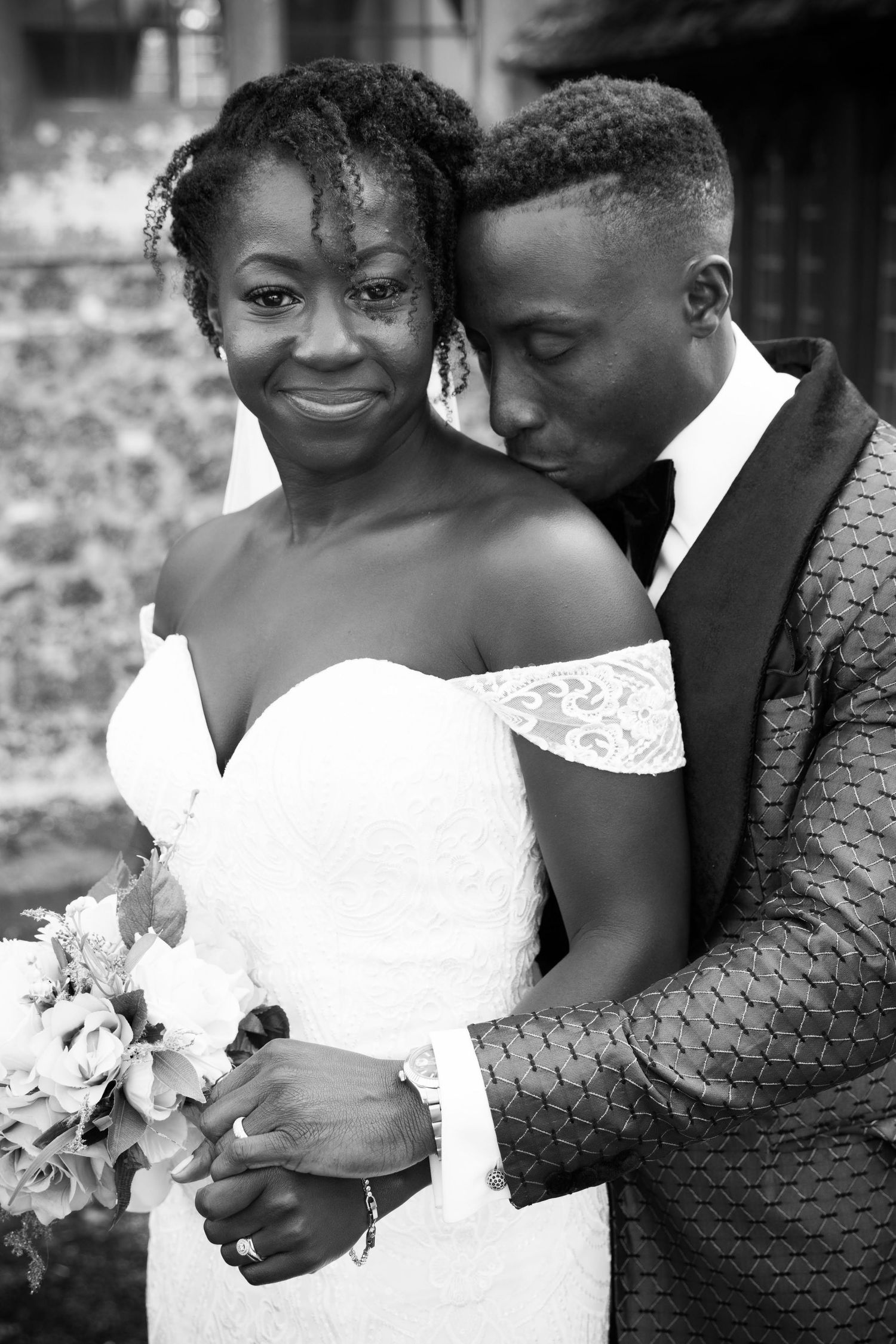 Black and white portrait of a smiling bride and groom embracing tenderly outside a stone building.