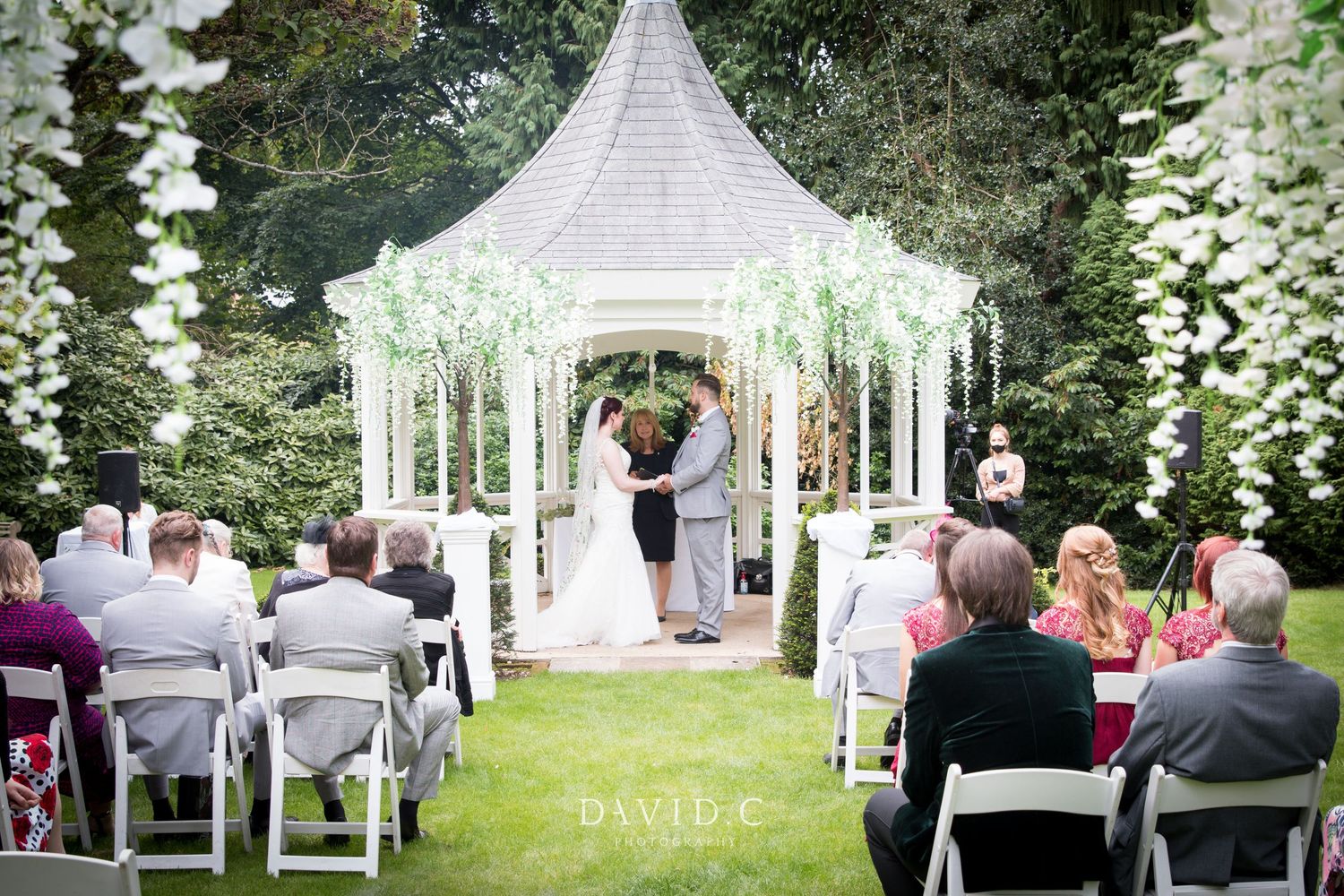 Wedding Photography - Outdoor wedding ceremony under a white gazebo adorned with flowers, guests seated on white chairs in a lush garden.