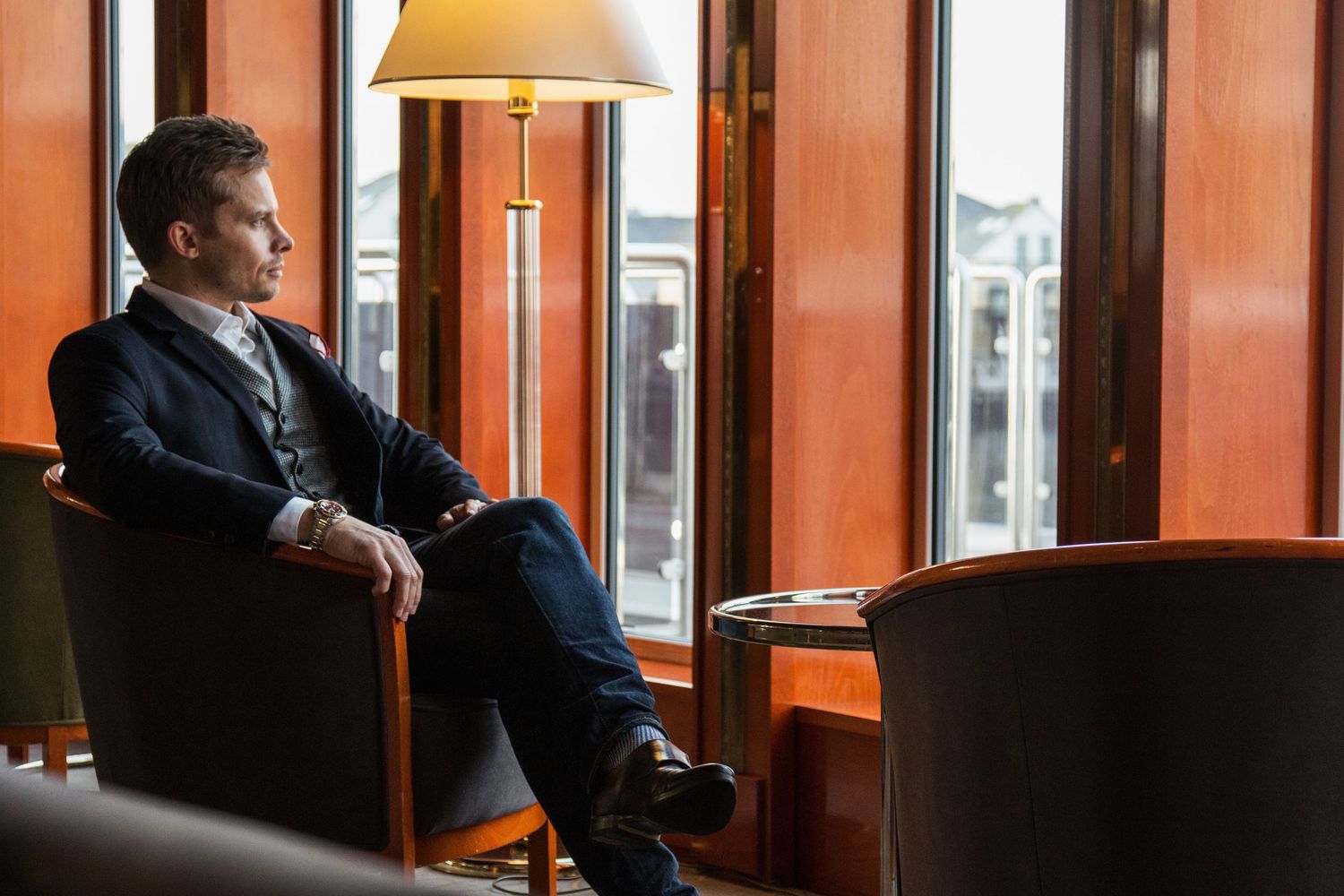 Portrait photography - Businessman in navy suit sitting in armchair by large windows in elegant hotel lounge.
