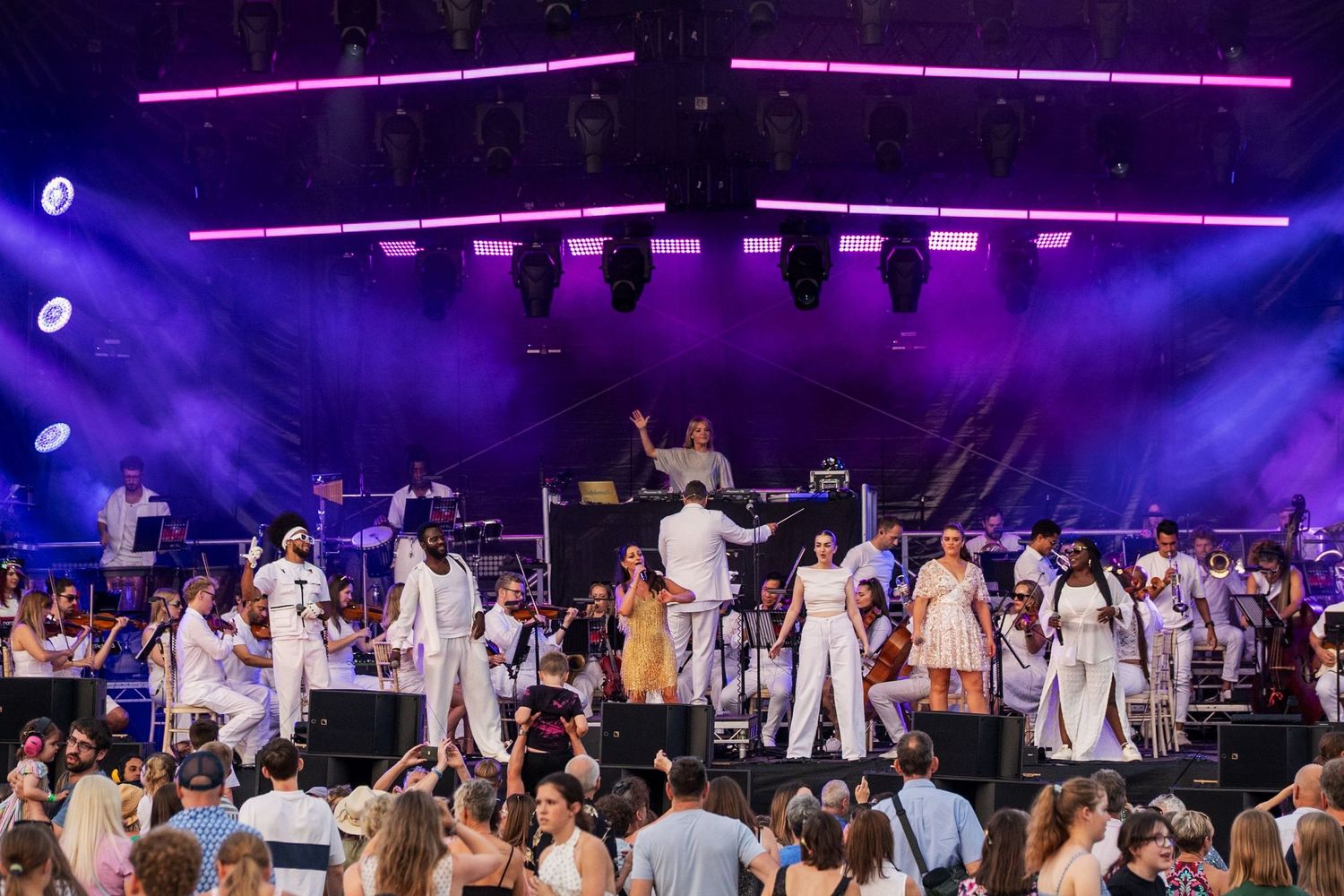 Large orchestra and choir performing on an outdoor festival stage with purple lighting and a conductor.