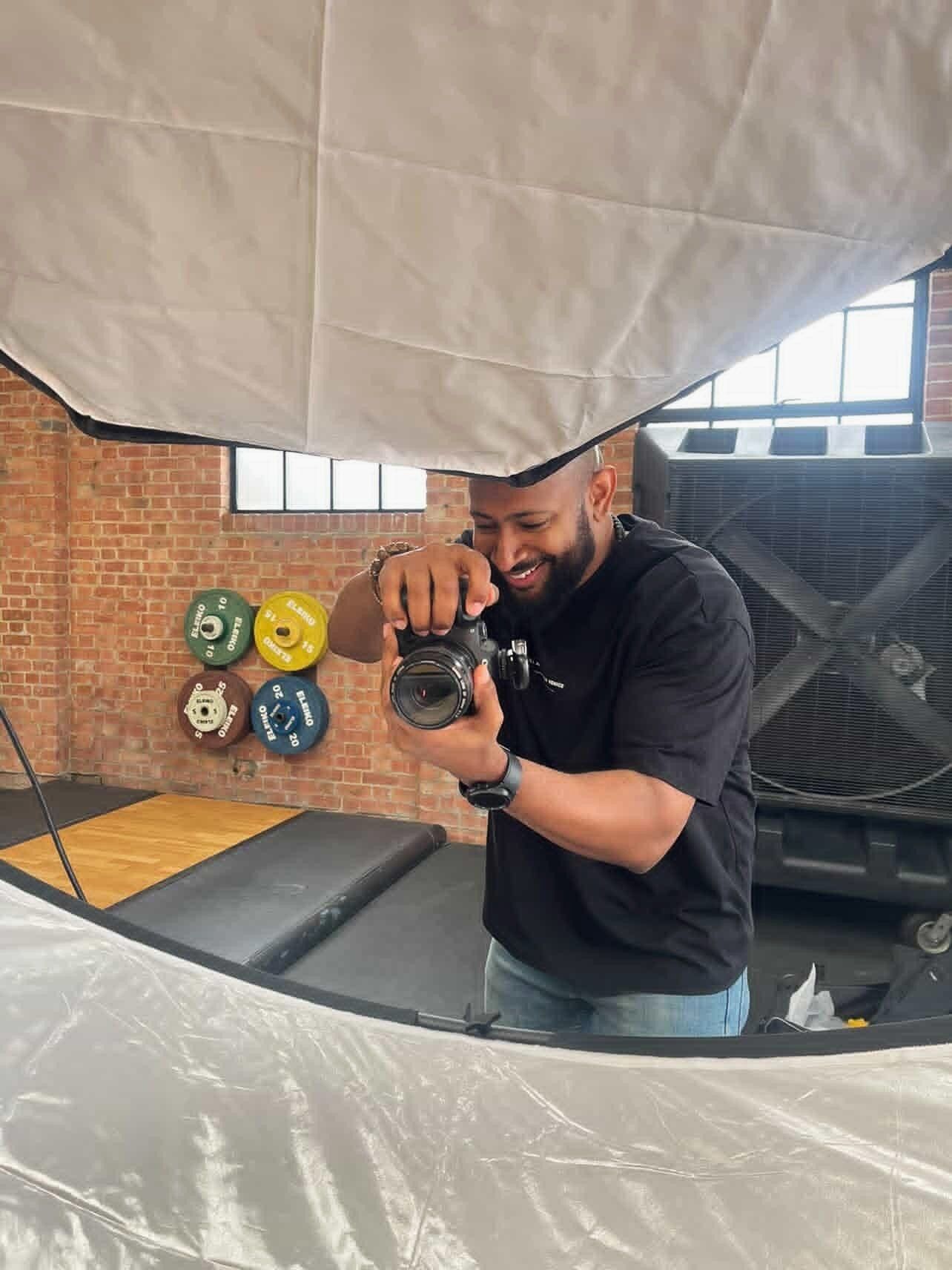 Smiling man in black t-shirt examining a camera lens inside a photography studio in a gym.