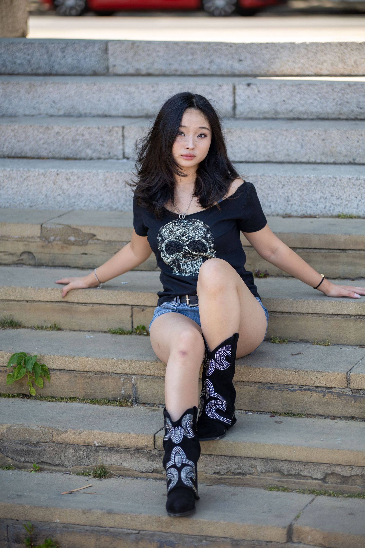 Portrait Photography - Young Asian woman in skull t-shirt, denim shorts, and cowboy boots sitting on stone steps.