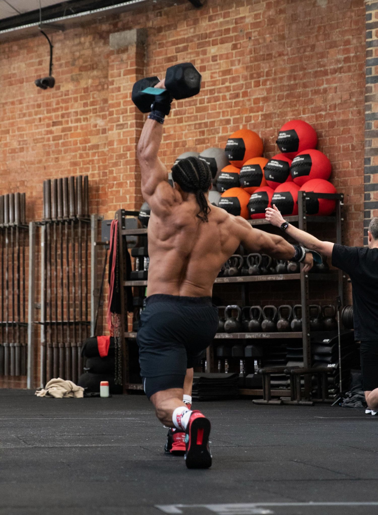 Fitness Photography - Shirtless muscular man performing a dumbbell snatch exercise in a CrossFit gym with kettlebells on rack.