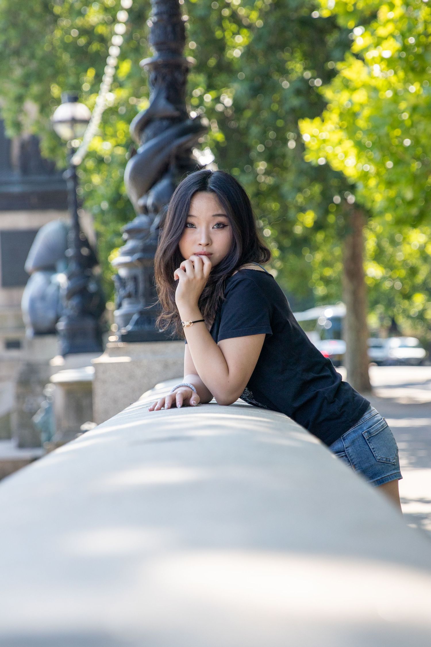 Young Asian woman in black t-shirt and denim shorts leaning on a wall in an urban park setting.