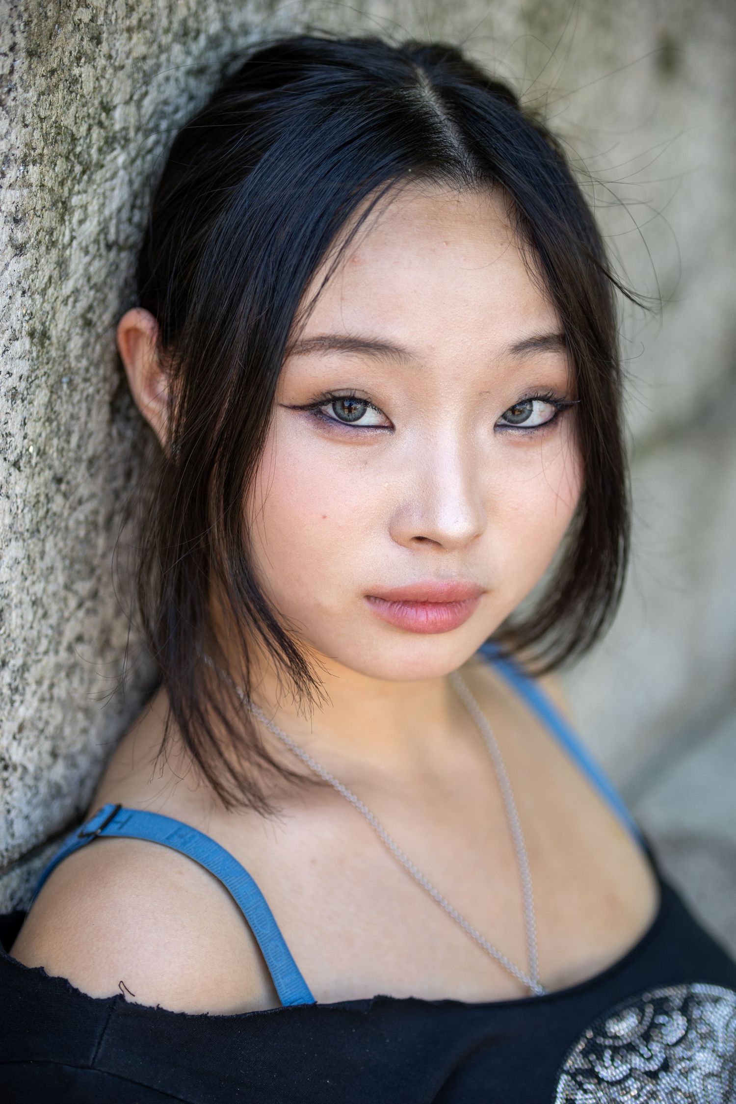 Close-up portrait of a young Asian woman with dark hair leaning against a stone wall.