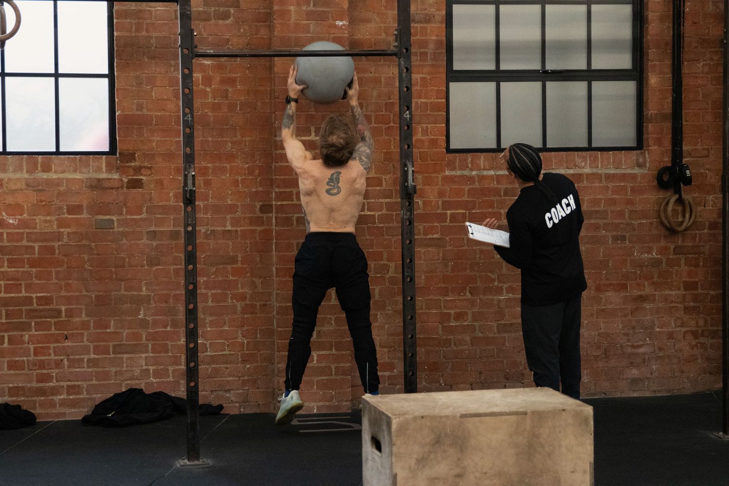 Shirtless athlete performing wall ball shots while a judge observes in a CrossFit gym.