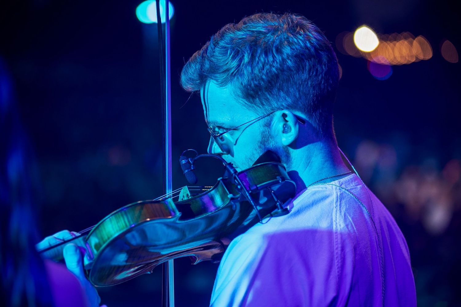 Male violinist wearing glasses performing on stage under blue and purple stage lighting at a concert.