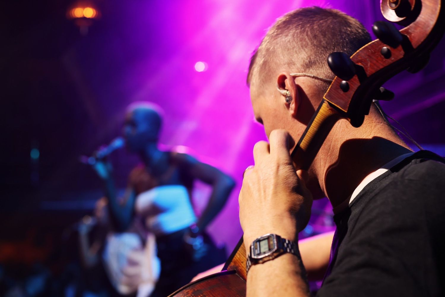 Live Music Event Photography - Cellist performing in the foreground at a live concert with a singer visible under pink stage lights.