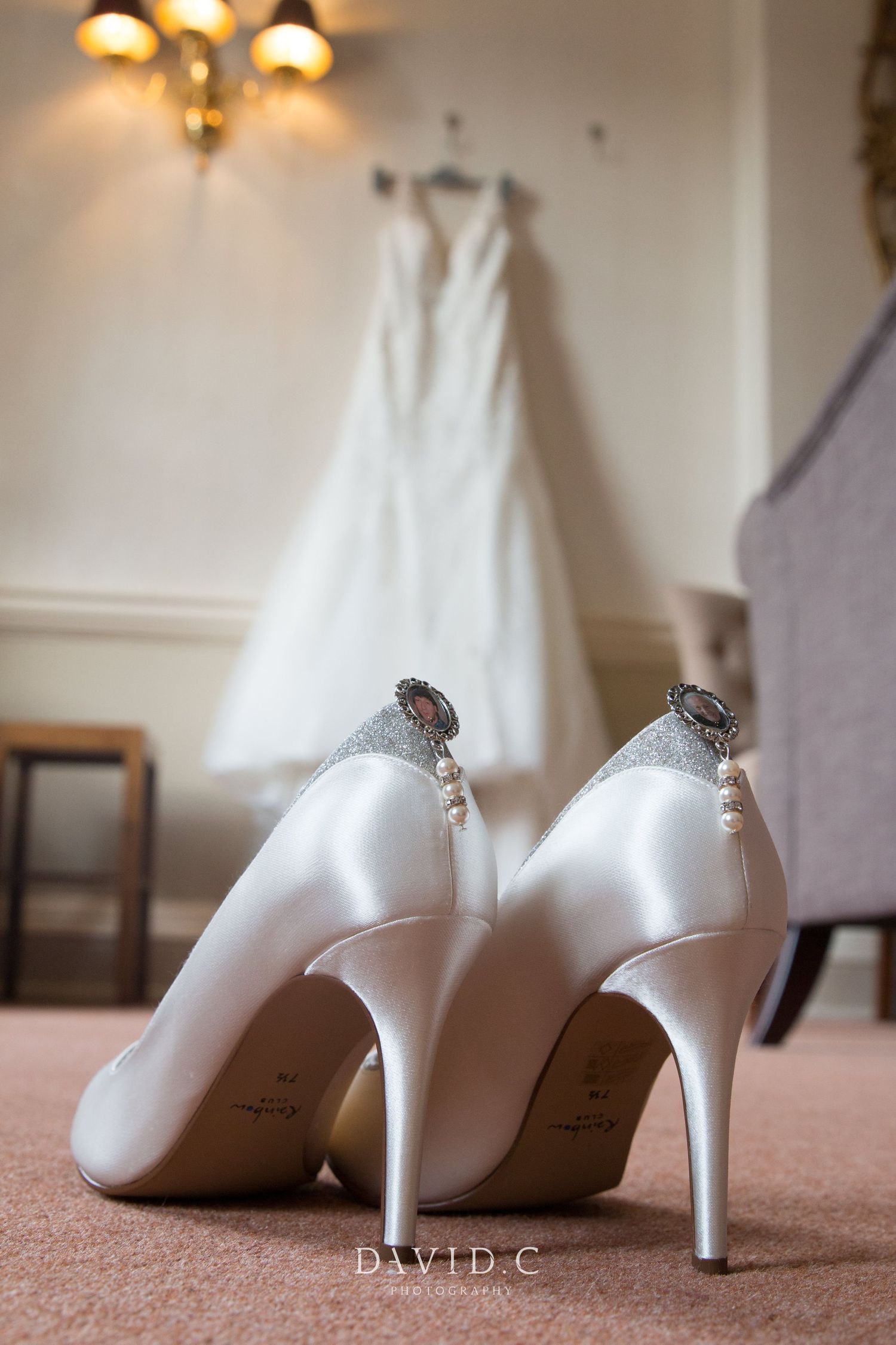 Wedding Photography - White satin bridal heels with pearl and brooch details displayed on carpet with wedding dress hanging in background.