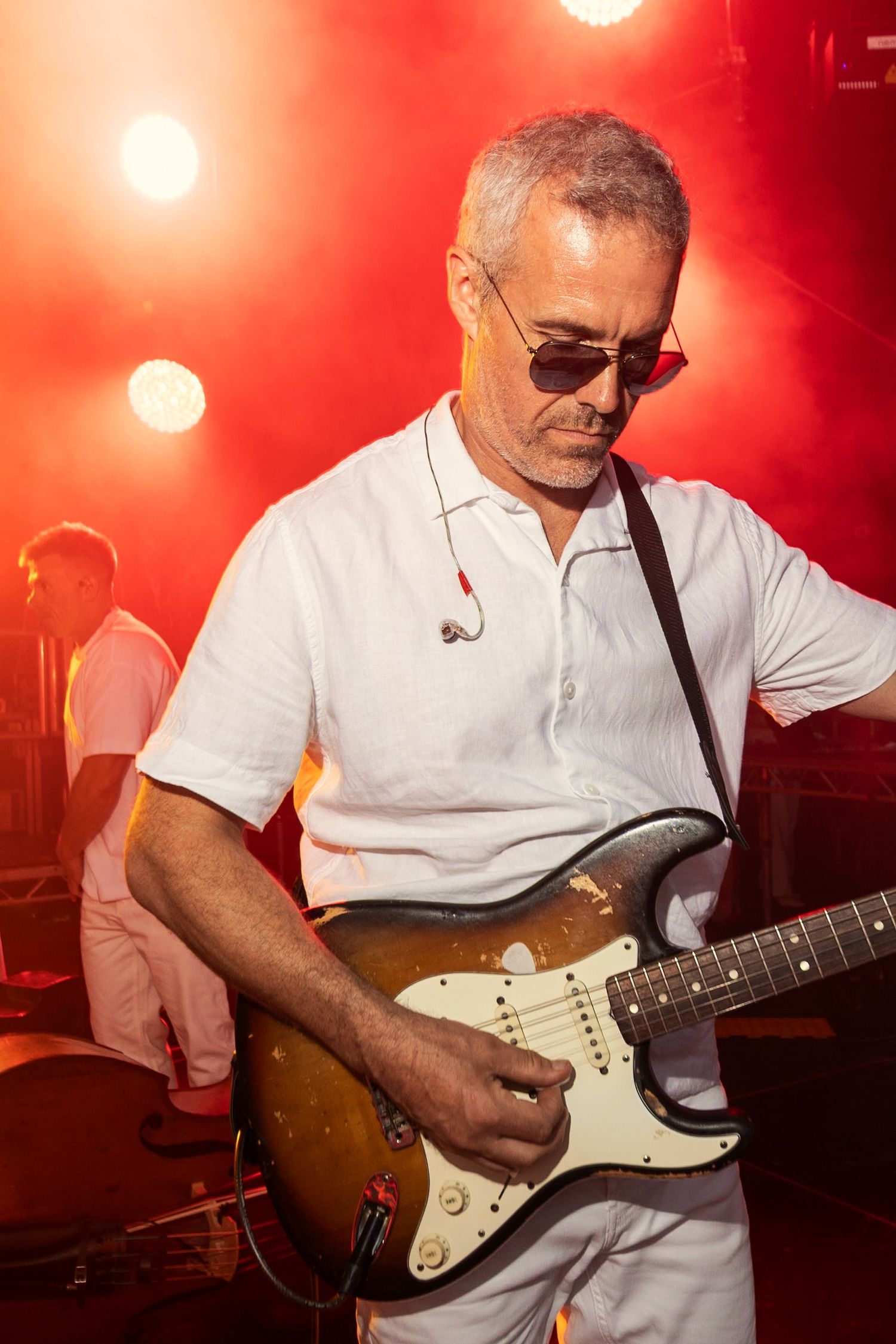 Male guitarist in white shirt and sunglasses playing a Fender Stratocaster on a red-lit concert stage.