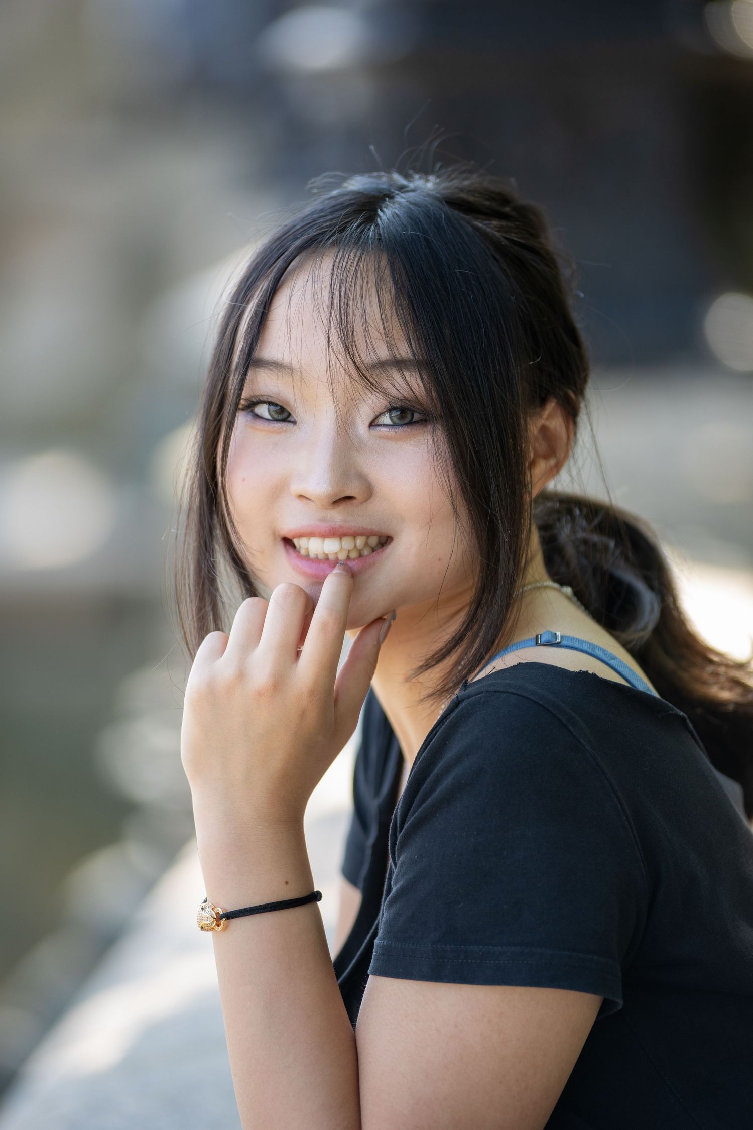 Smiling young Asian woman in black t-shirt touching her lips near a sunlit water feature.