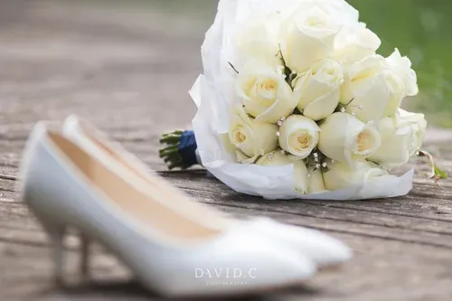 Wedding Photography - White bridal heels and a bouquet of white roses resting on a rustic wooden surface outdoors.