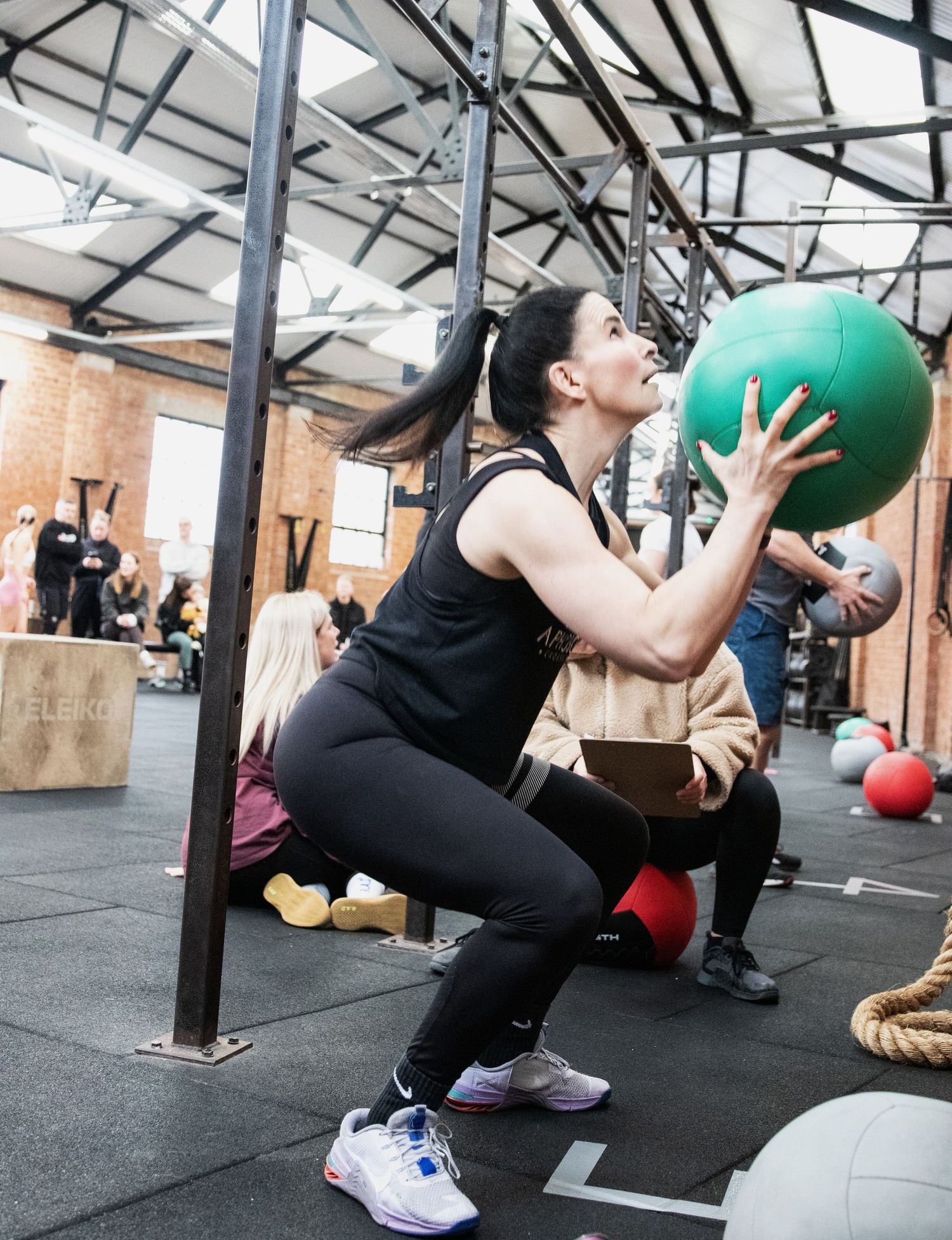 Woman doing a wall ball squat exercise with green medicine ball in a busy CrossFit gym.