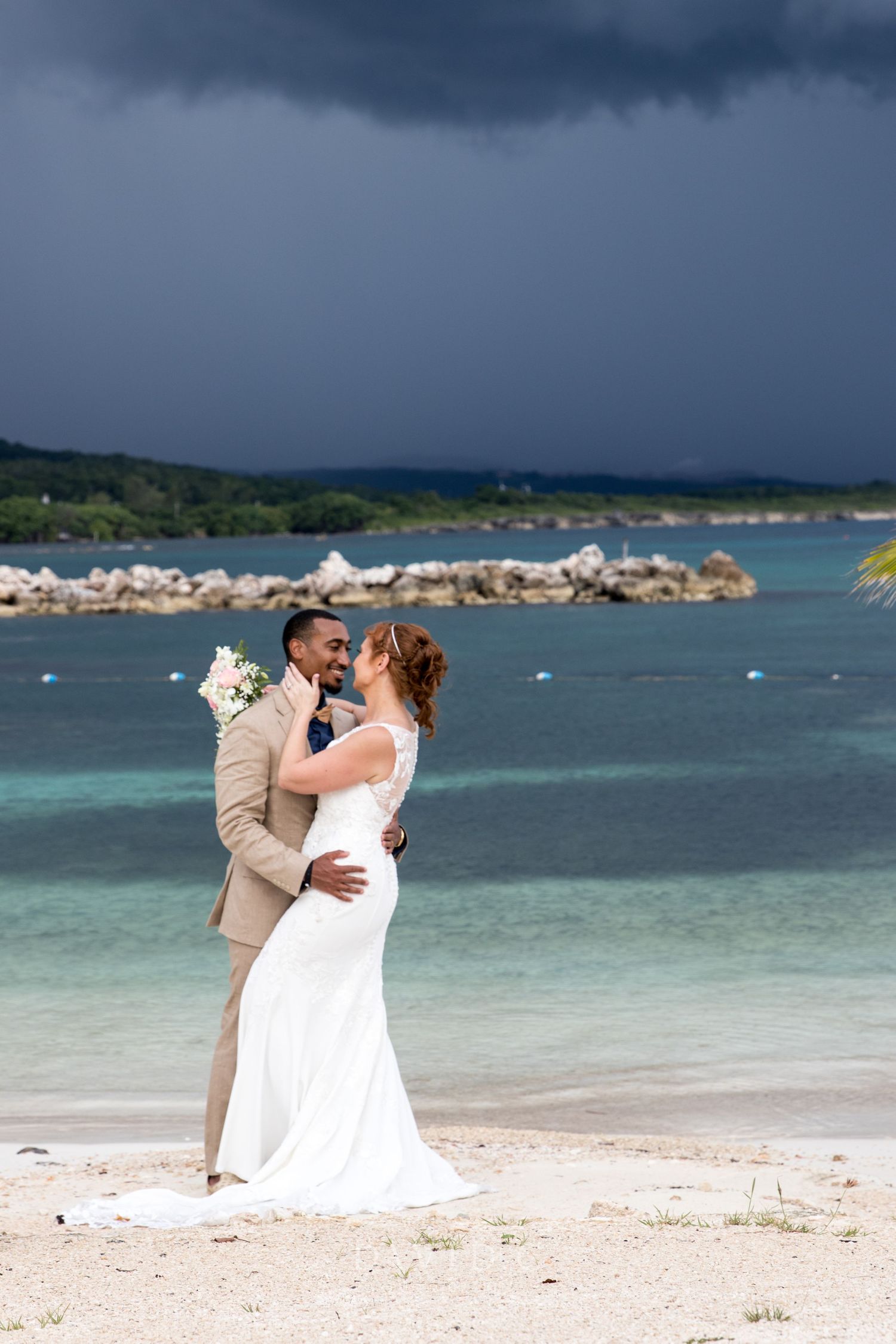 Wedding Photography - Interracial couple in wedding attire embracing on a tropical beach with turquoise water and a stormy sky.