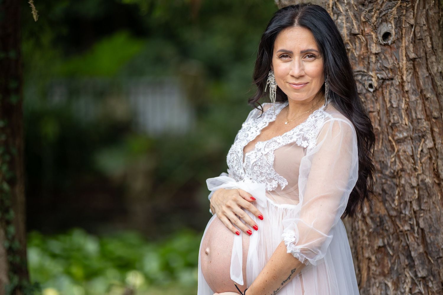 Portrait photography - Smiling pregnant woman in sheer lace maternity robe cradling baby bump against tree outdoors.