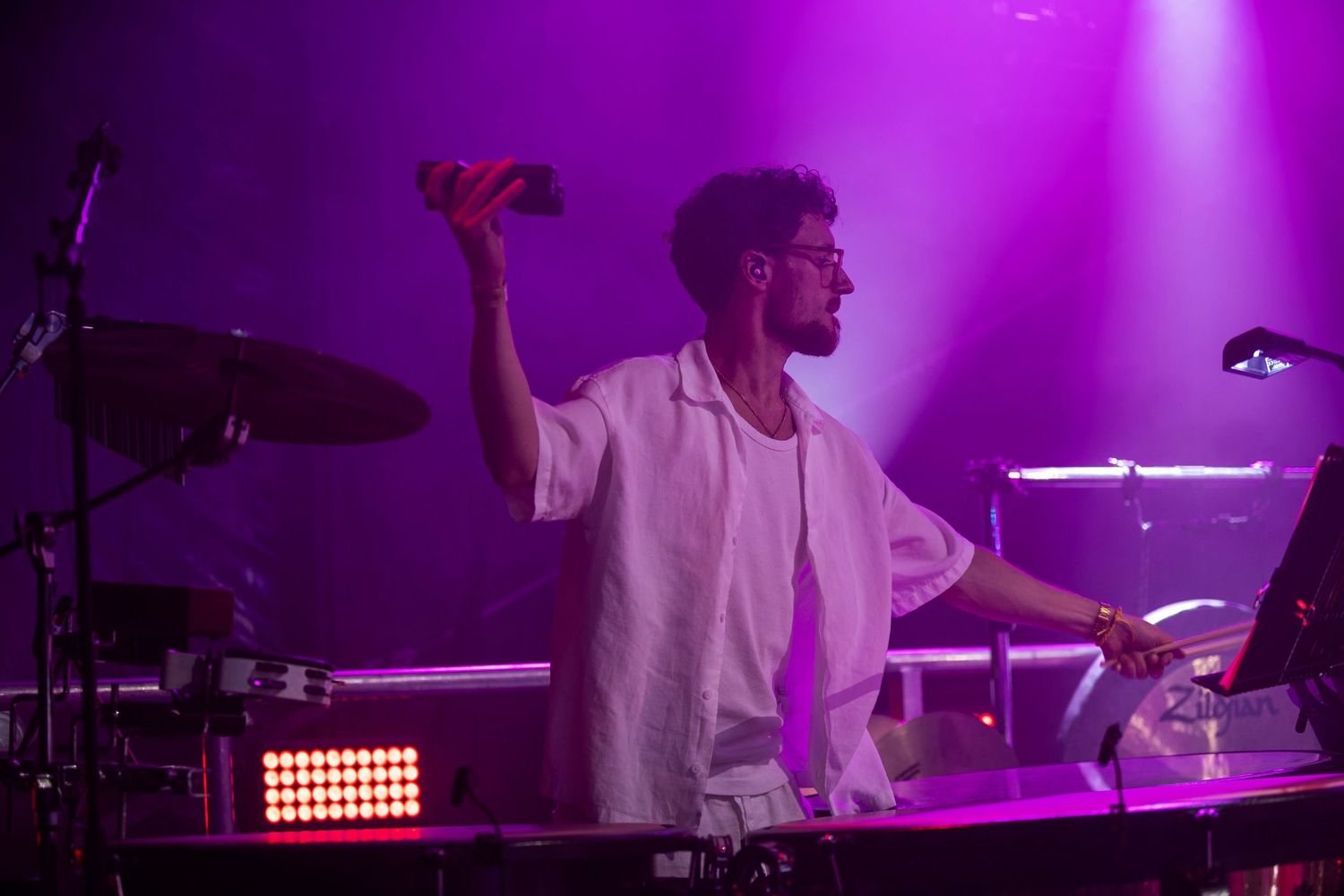 Male performer raises his hand on stage bathed in purple light while playing drums at a live concert.