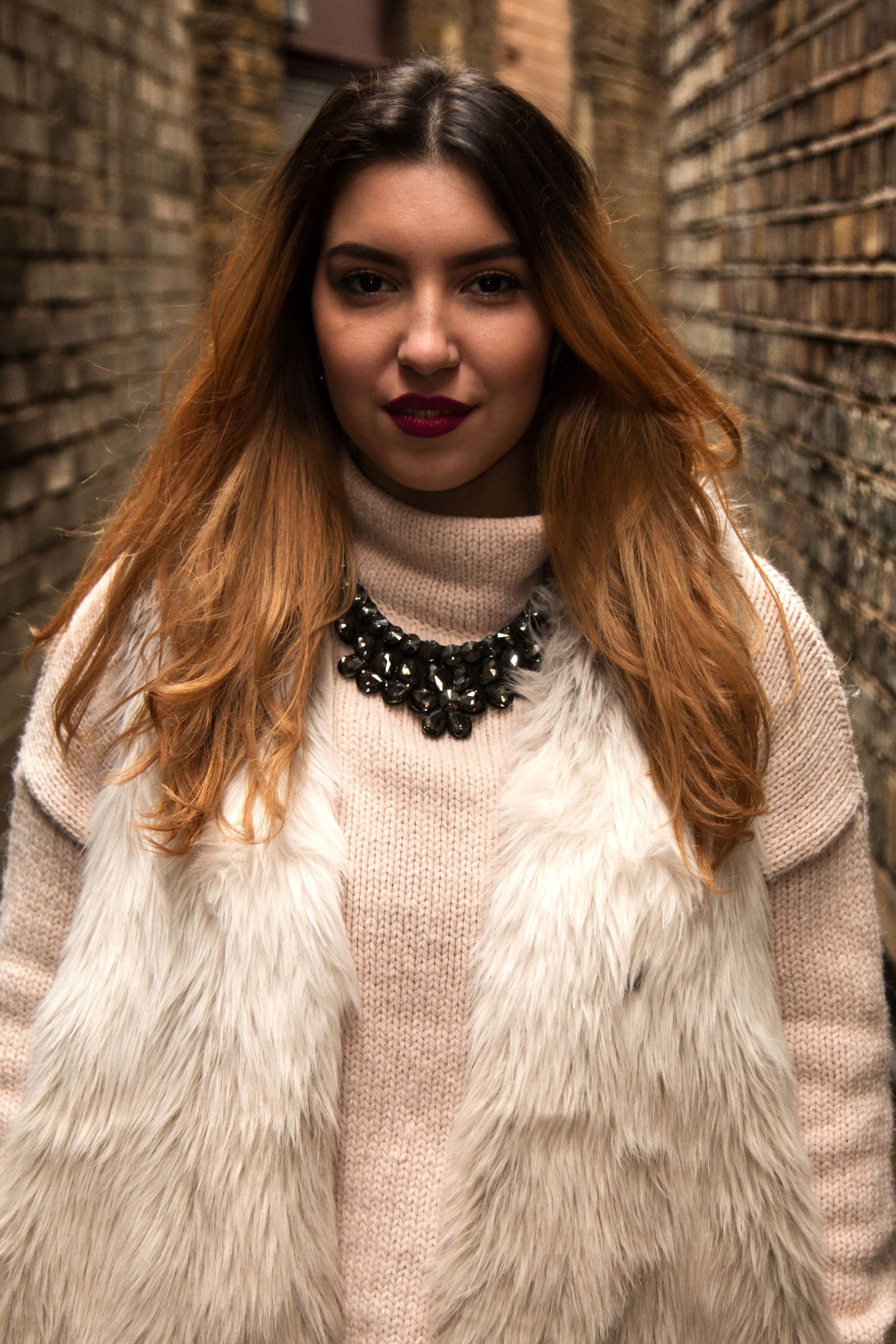 Portrait photography - Stylish young woman in fur vest and pink sweater with statement necklace in urban alleyway.