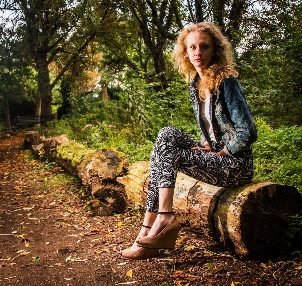 Portrait photography - Young woman with curly hair sitting on fallen logs in autumn forest wearing denim jacket.