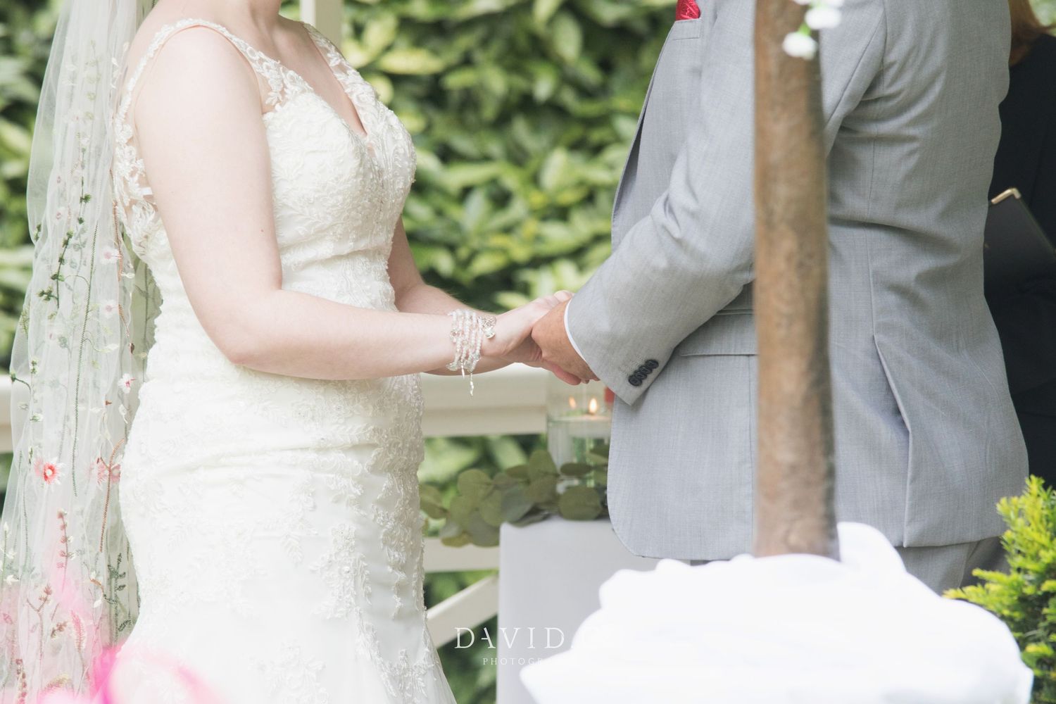 Bride in lace gown and groom in grey suit holding hands during outdoor garden wedding ceremony.