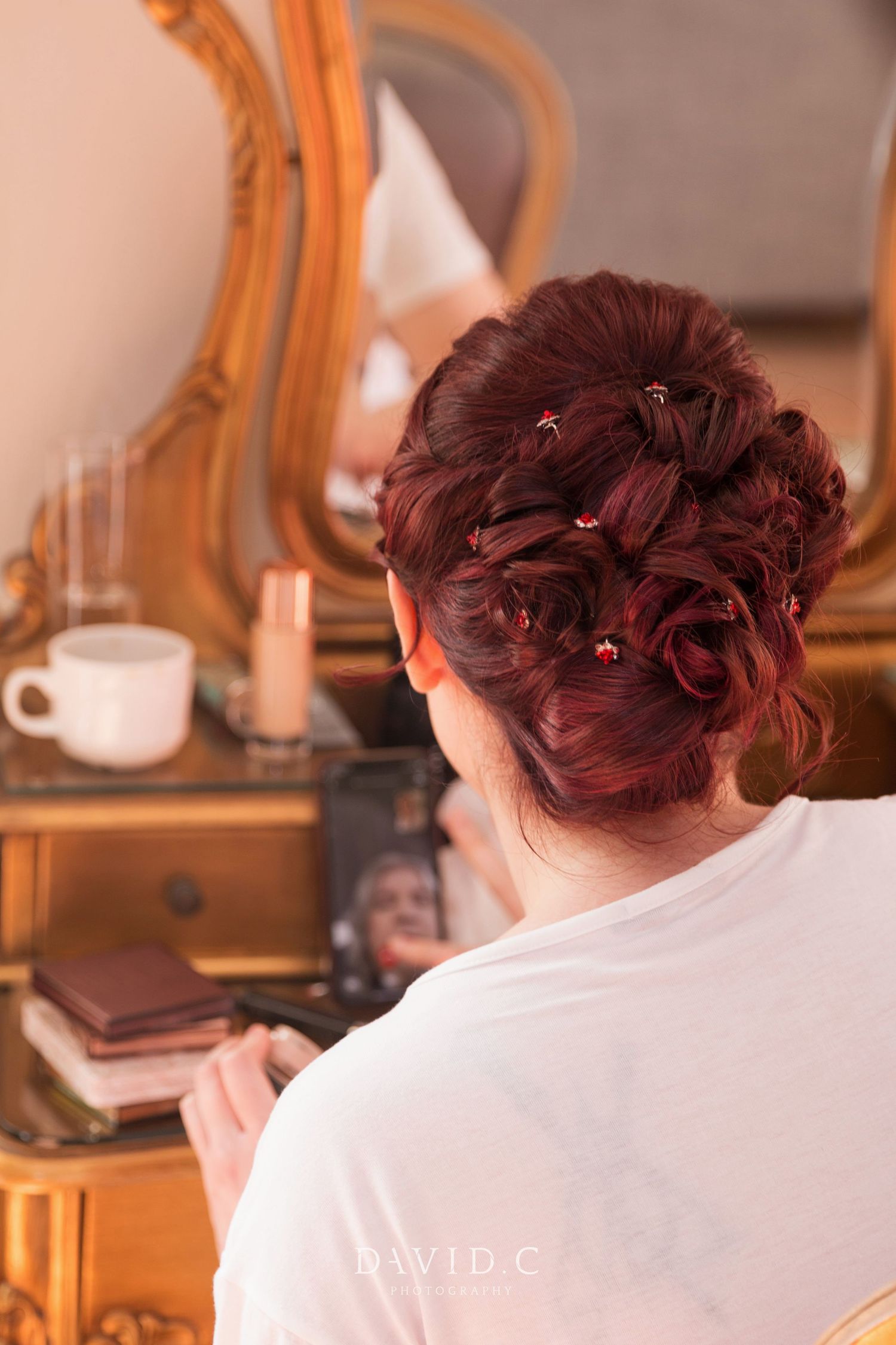 Bride with elegant red updo hairstyle adorned with small floral pins sitting at vintage gold mirror vanity.