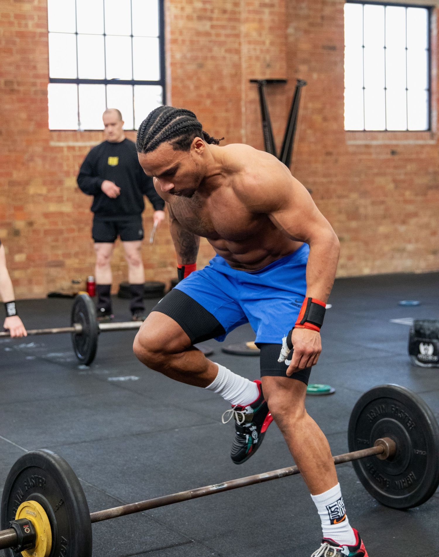 Shirtless male athlete preparing to lift a barbell in a brick-walled CrossFit gym.