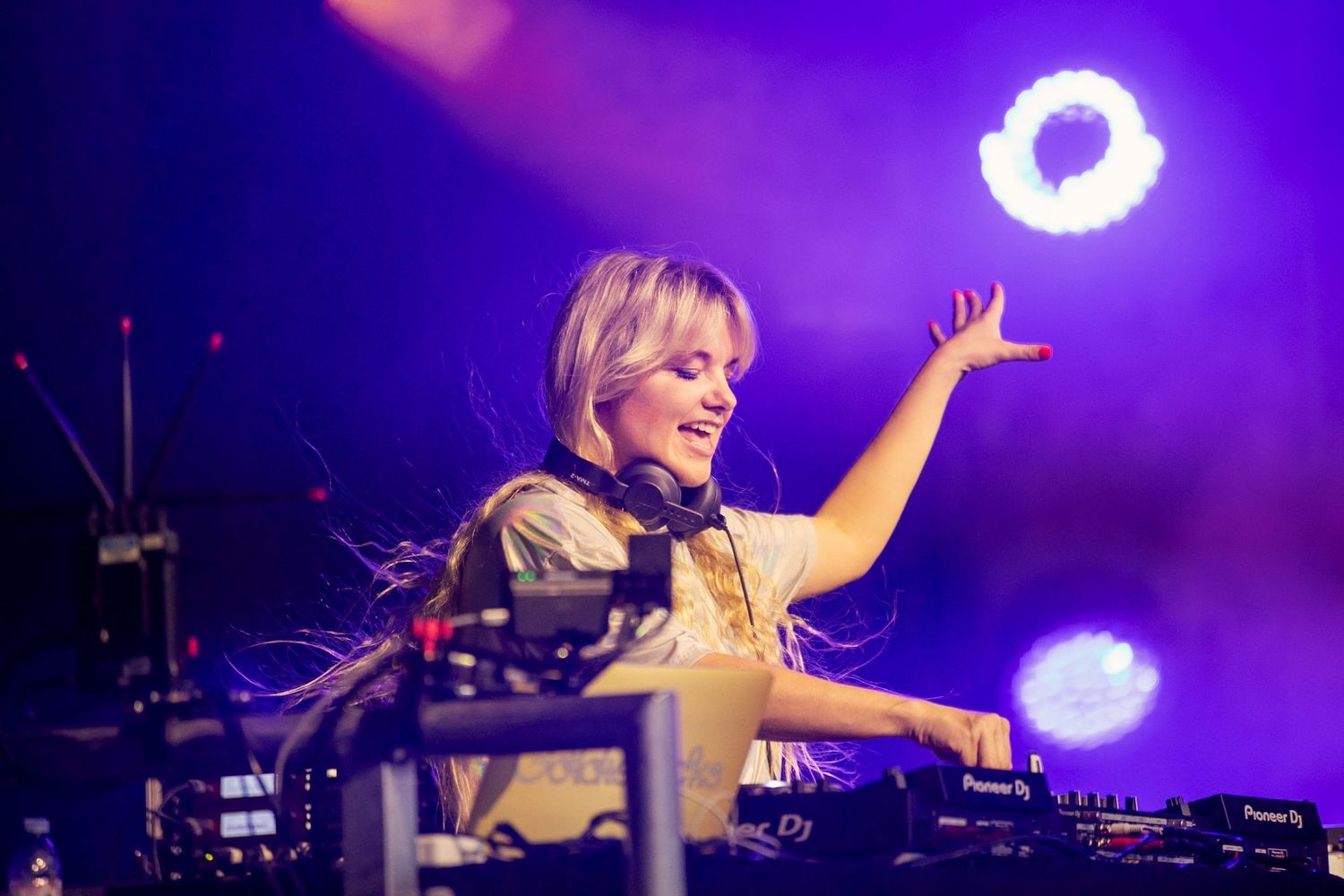 Female DJ performing at a live music festival, smiling and raising her hand with DJ equipment and colorful stage lights.
