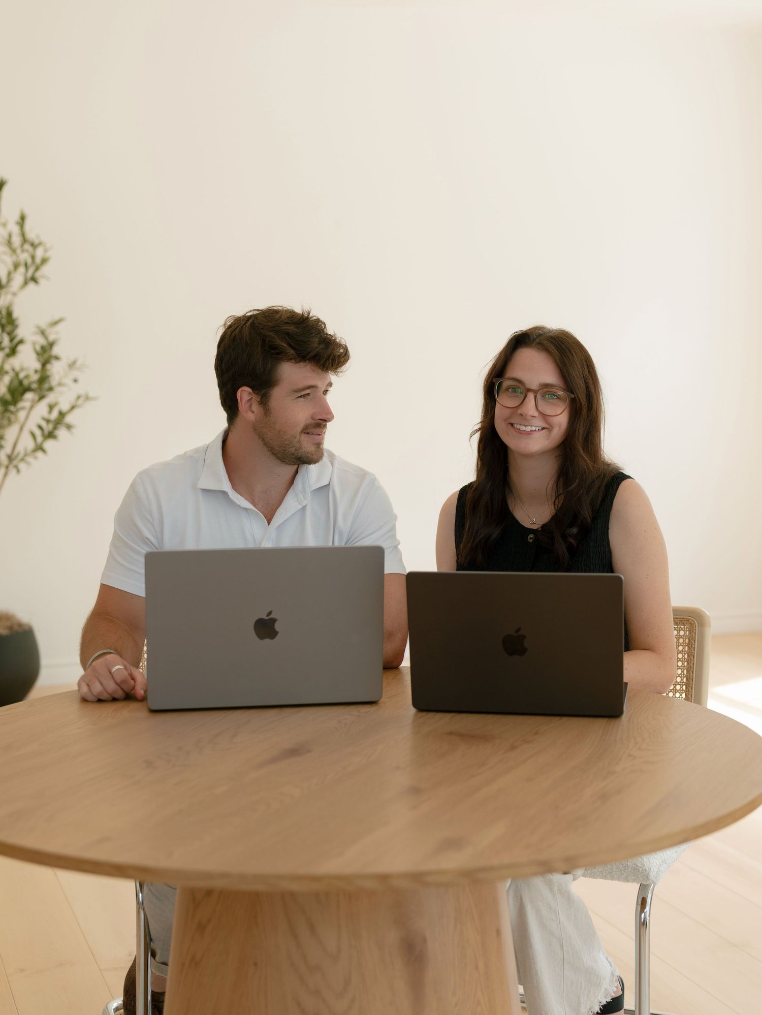Two professionals smiling and working on Apple MacBooks at a round wooden table in a bright modern office.