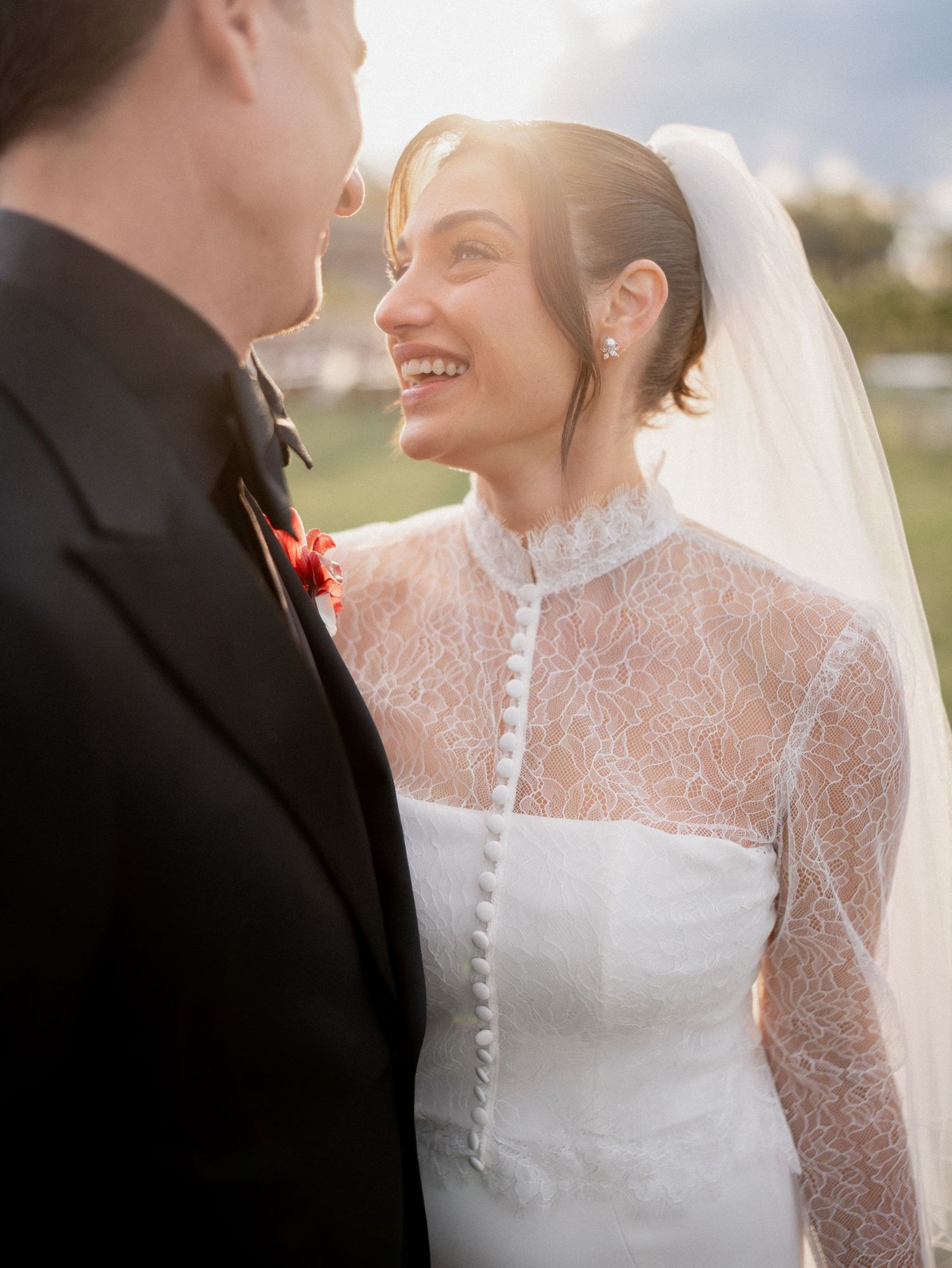Bride in lace high-neck wedding gown and veil smiles at groom in black tuxedo with red boutonniere outdoors.