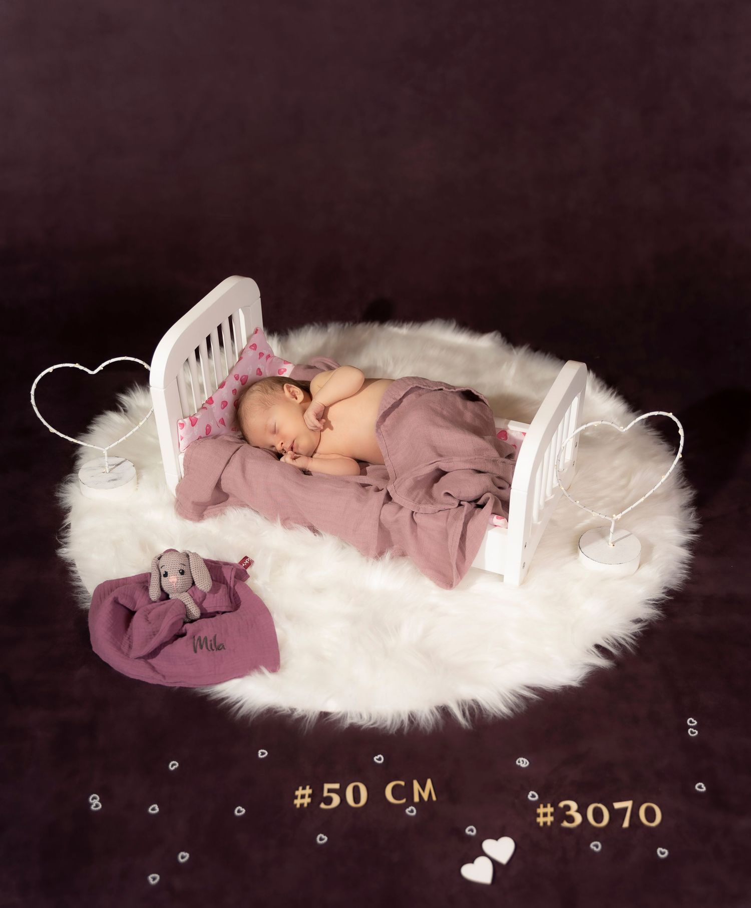 Newborn baby sleeping peacefully in a tiny white crib on fluffy white rug with purple blanket and stuffed animal toy.