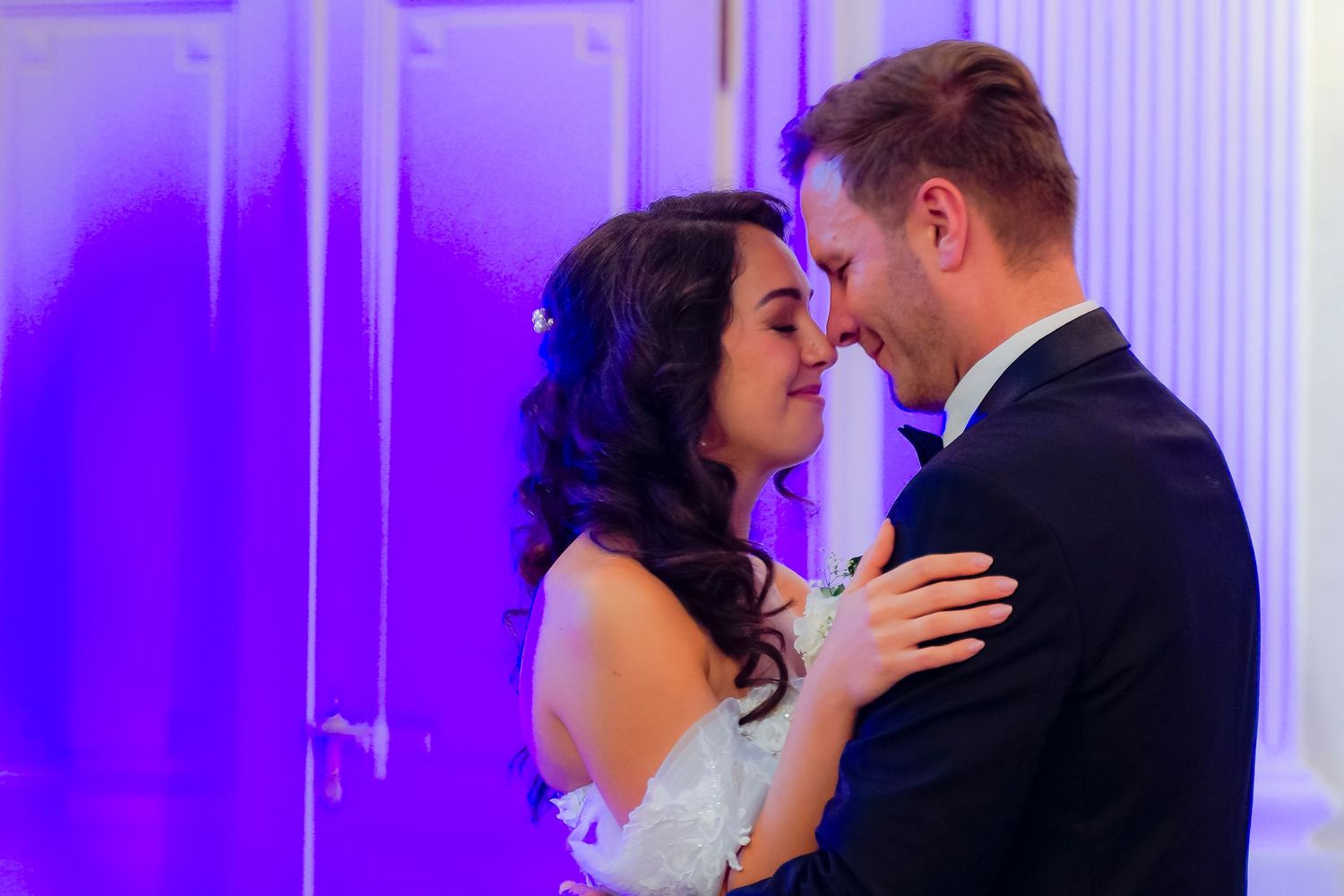 Bride and groom share a tender moment, foreheads touching, during their first dance in a ballroom with soft purple lighting.