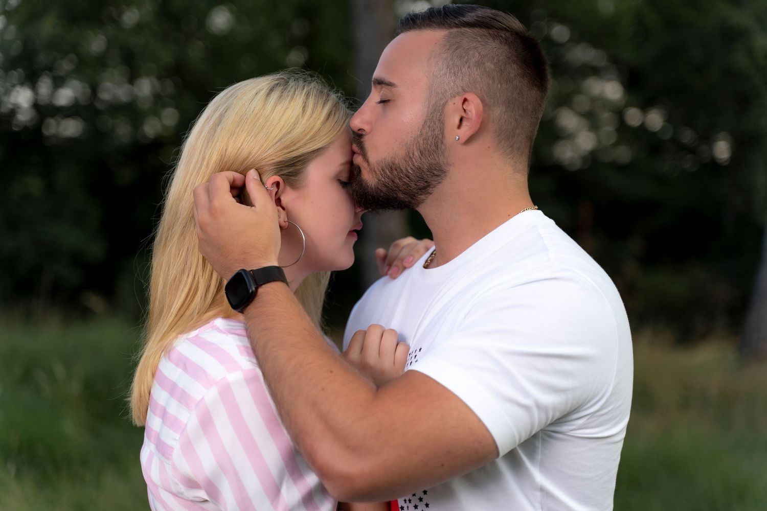 A couple sharing a romantic kiss outdoors, with the man in a white shirt embracing a blonde woman in a pink top.