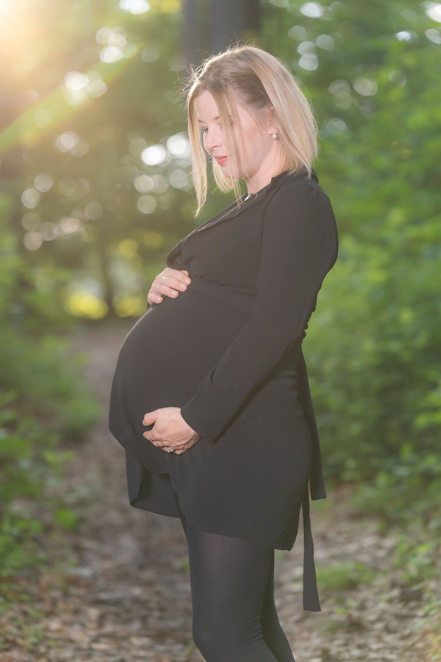 Pregnant woman in black dress cradling her baby bump outdoors in a sunlit forest path.