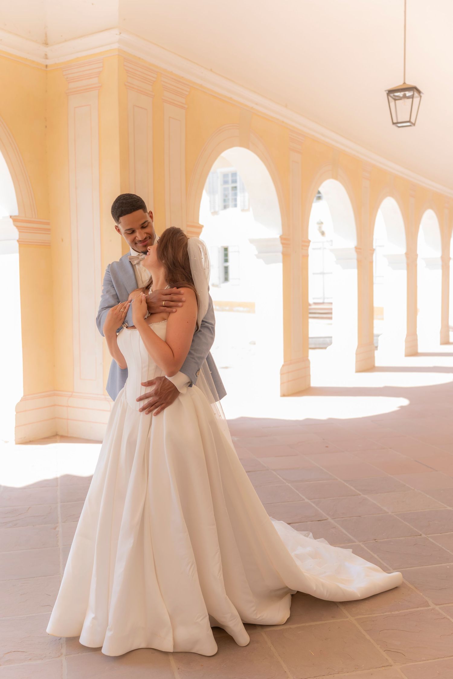Bride in a white gown embraces her groom in a gray suit beneath sunlit arched columns on a terracotta terrace.