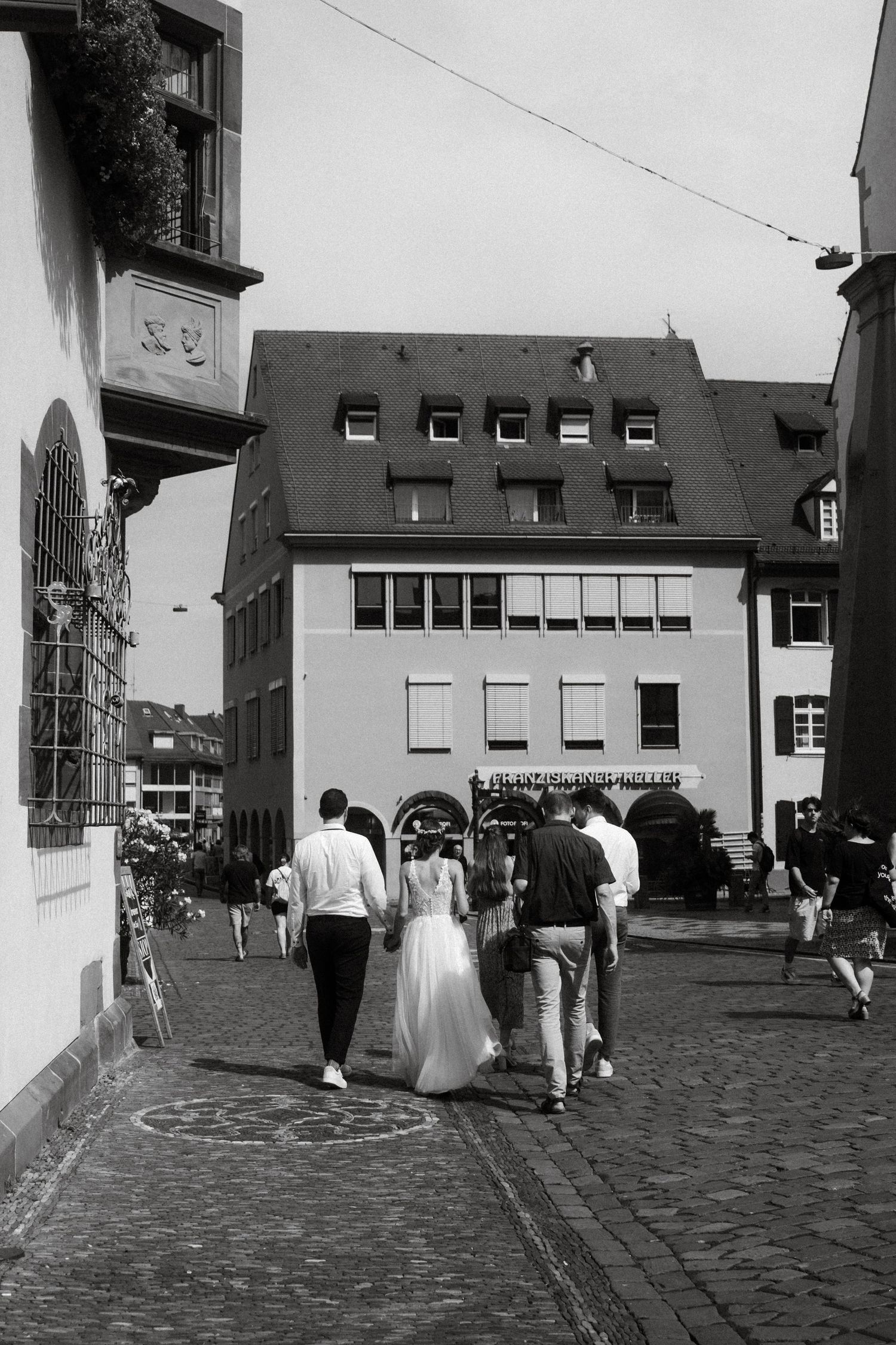 Newlyweds and guests walking through a city with buildings and cobblestones.